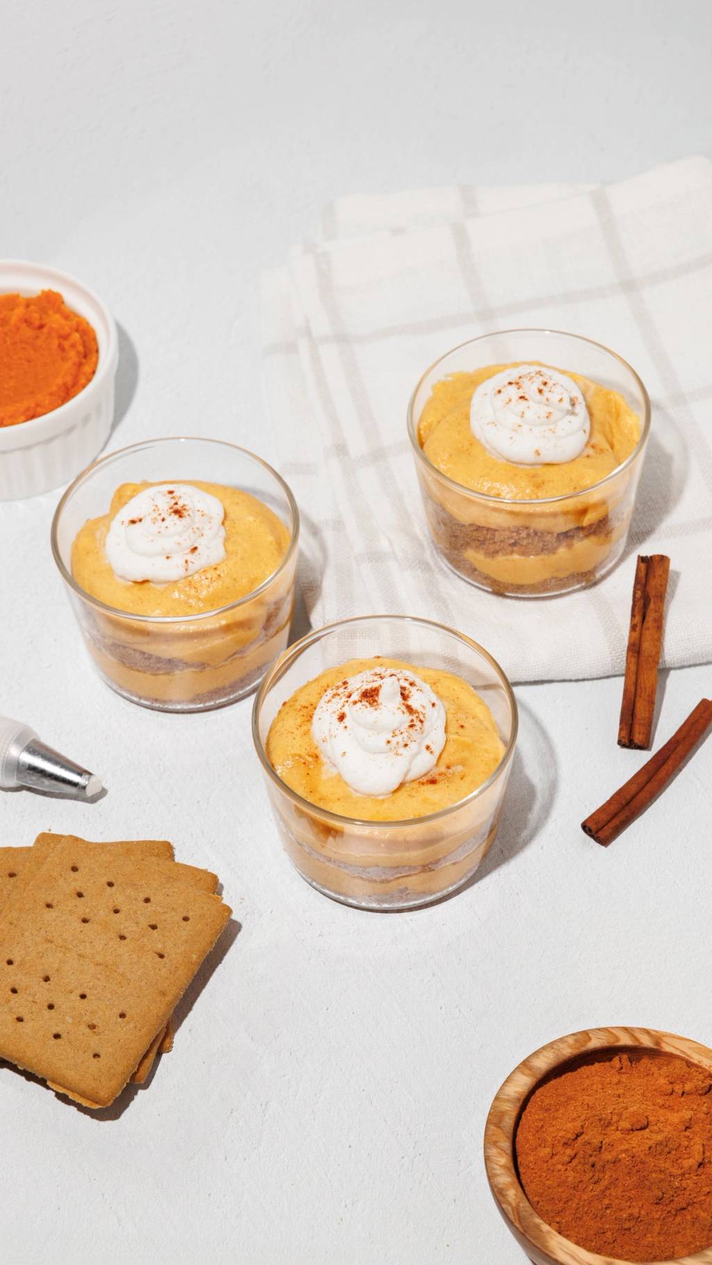 Three layered orange desserts topped with whipped cream, with crackers, cinnamon sticks, and bowls of ingredients nearby.