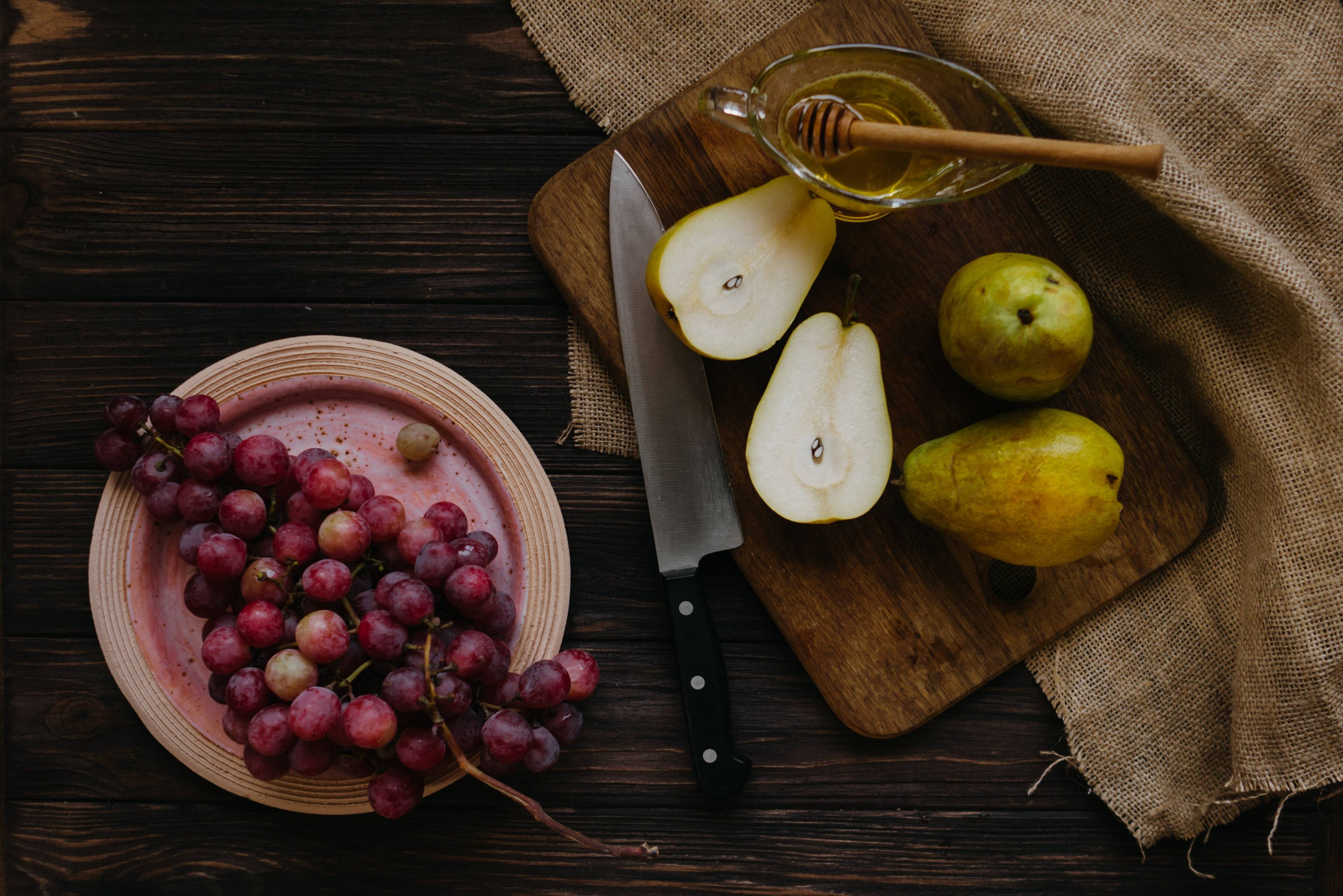 Red grapes, whole and halved pears, honey, and a knife on a wooden table