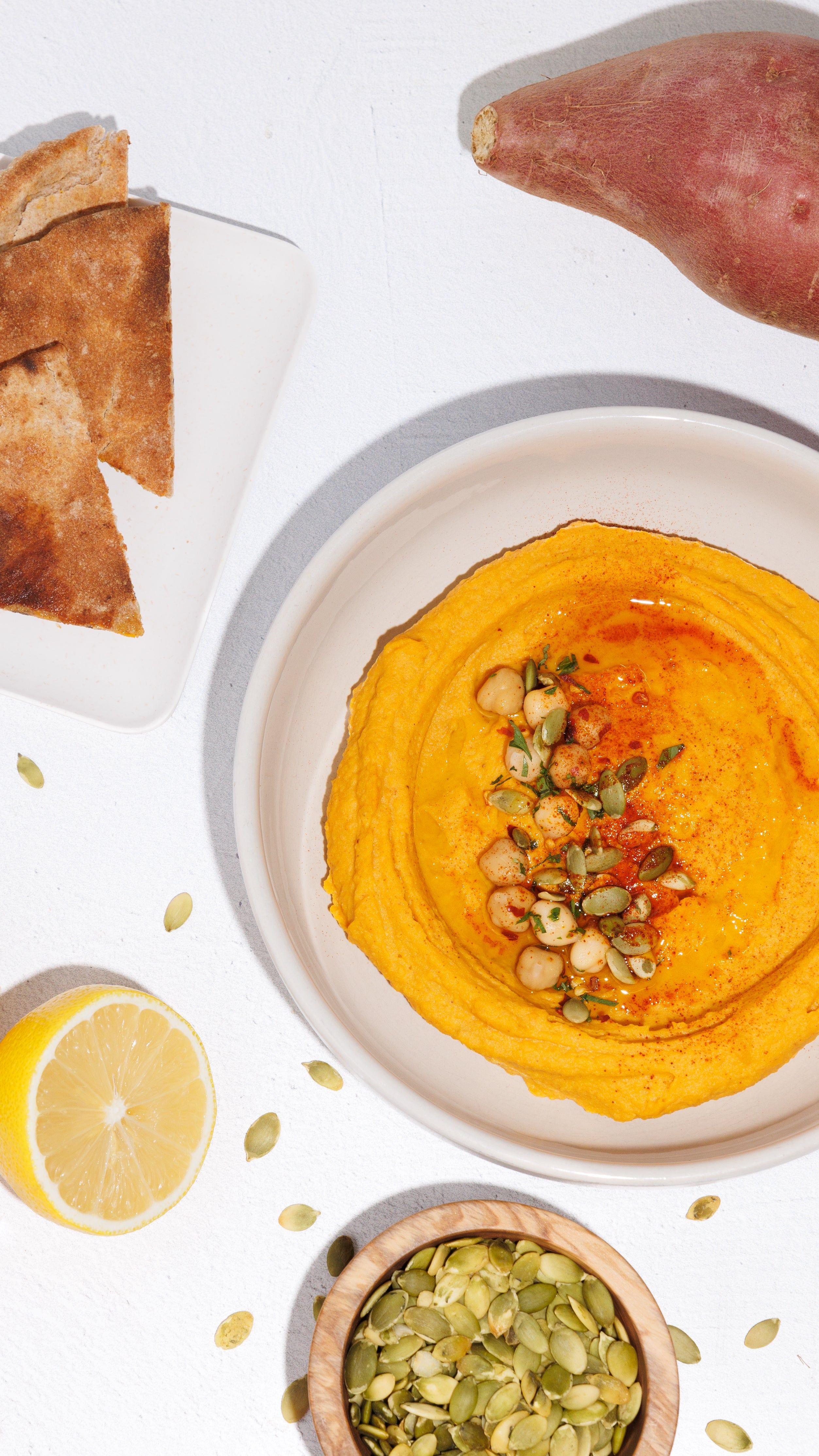 Bowl of orange sweet potato hummus topped with chickpeas and pumpkin seeds, with pita, lemon, and sweet potato.