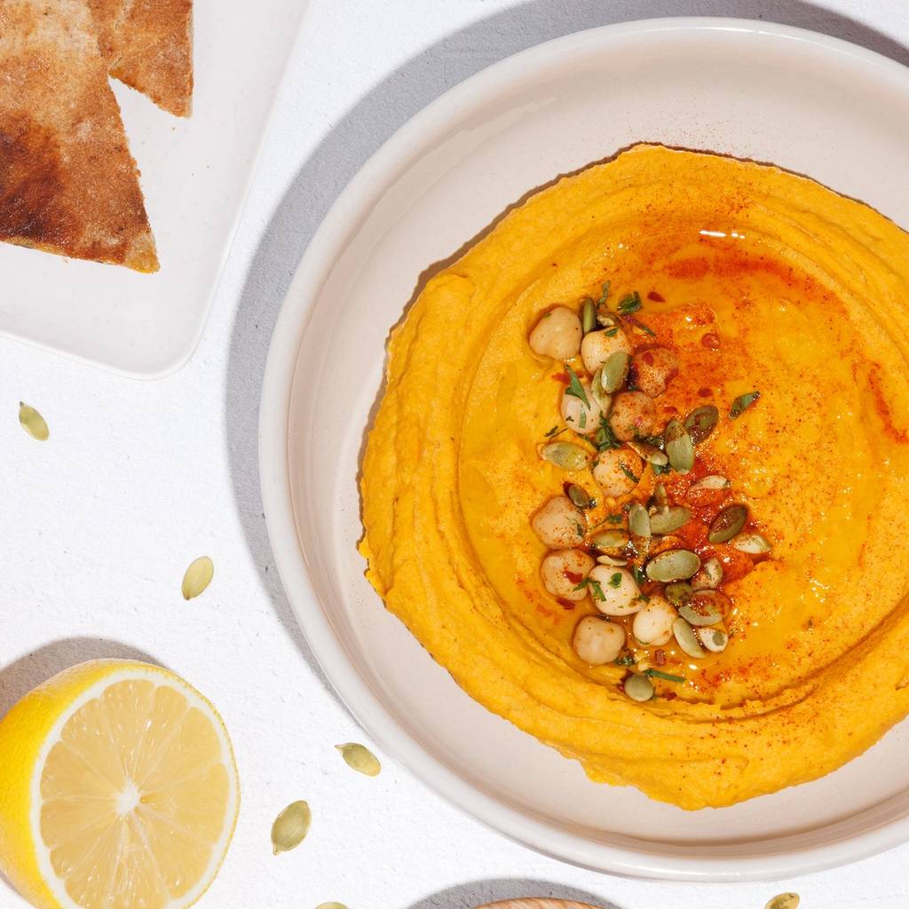 Bowl of orange sweet potato hummus topped with chickpeas and pumpkin seeds, with pita, lemon, and sweet potato.