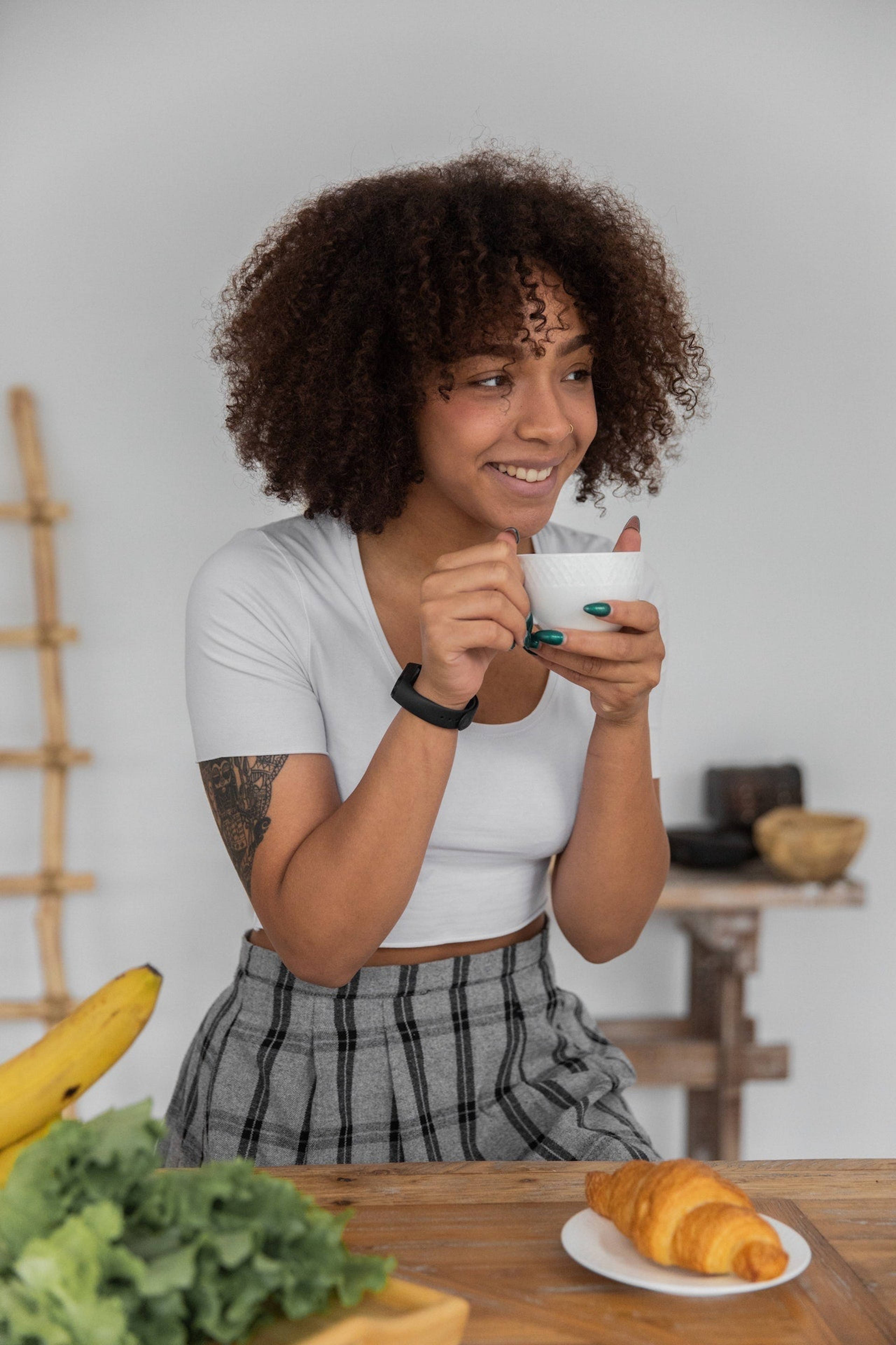 Smiling woman holding a white cup beside a croissant, banana, and leafy greens on a table.