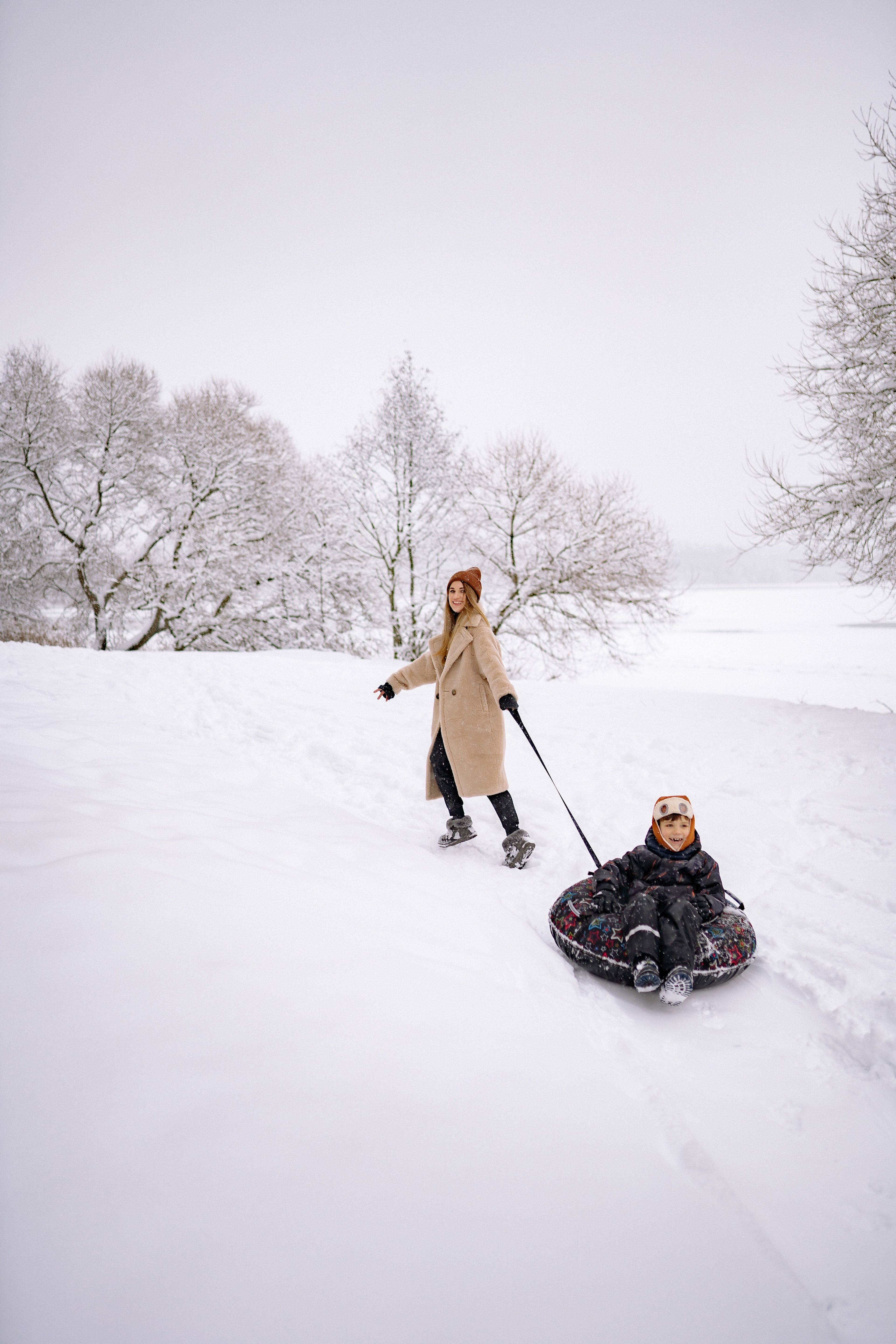 Woman in a tan coat pulls a smiling child on a snow tube through a snowy park.