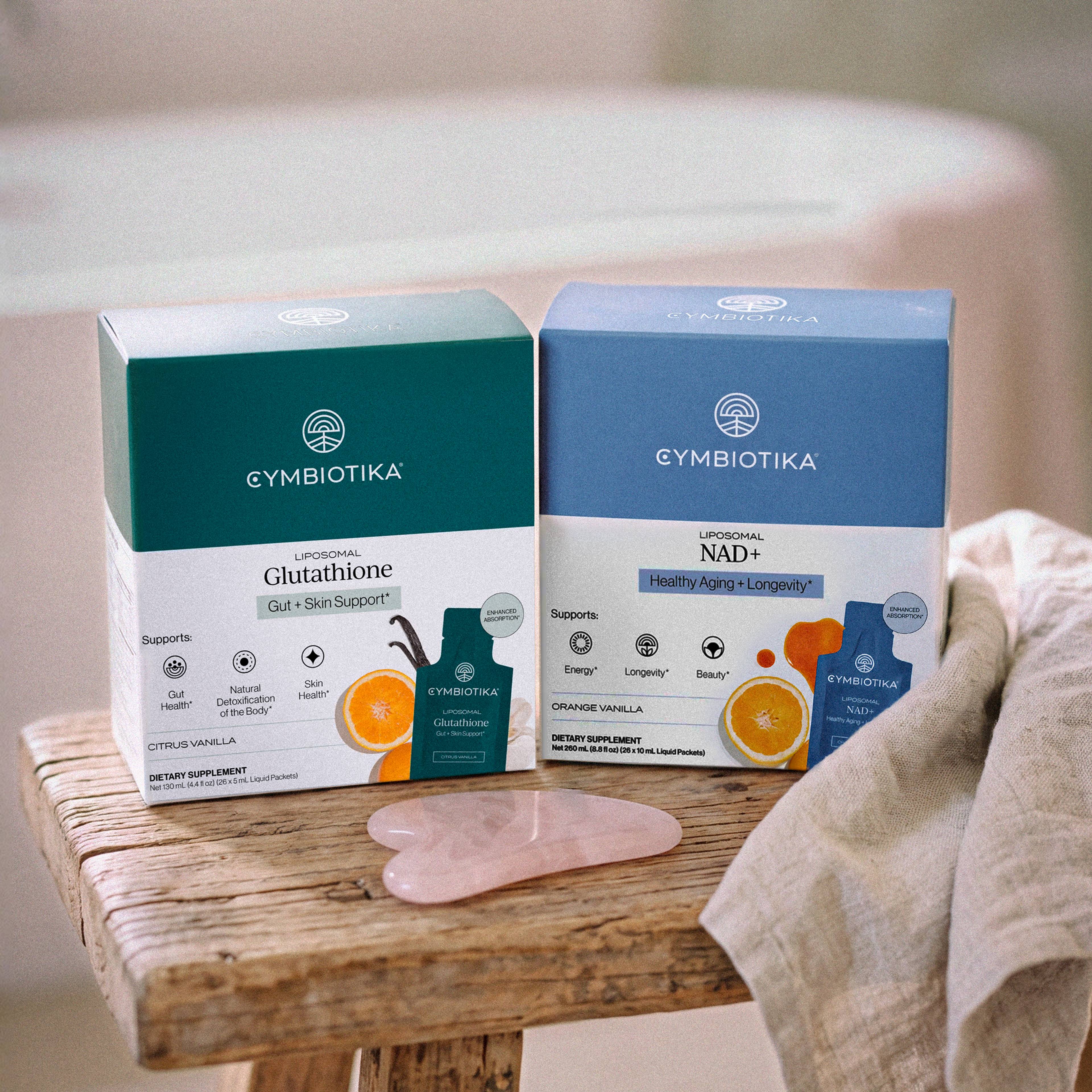 Cymbiotika Liposomal Glutathione and Liposomal NAD+ boxes beside a pink facial massage tool on a wooden stool.