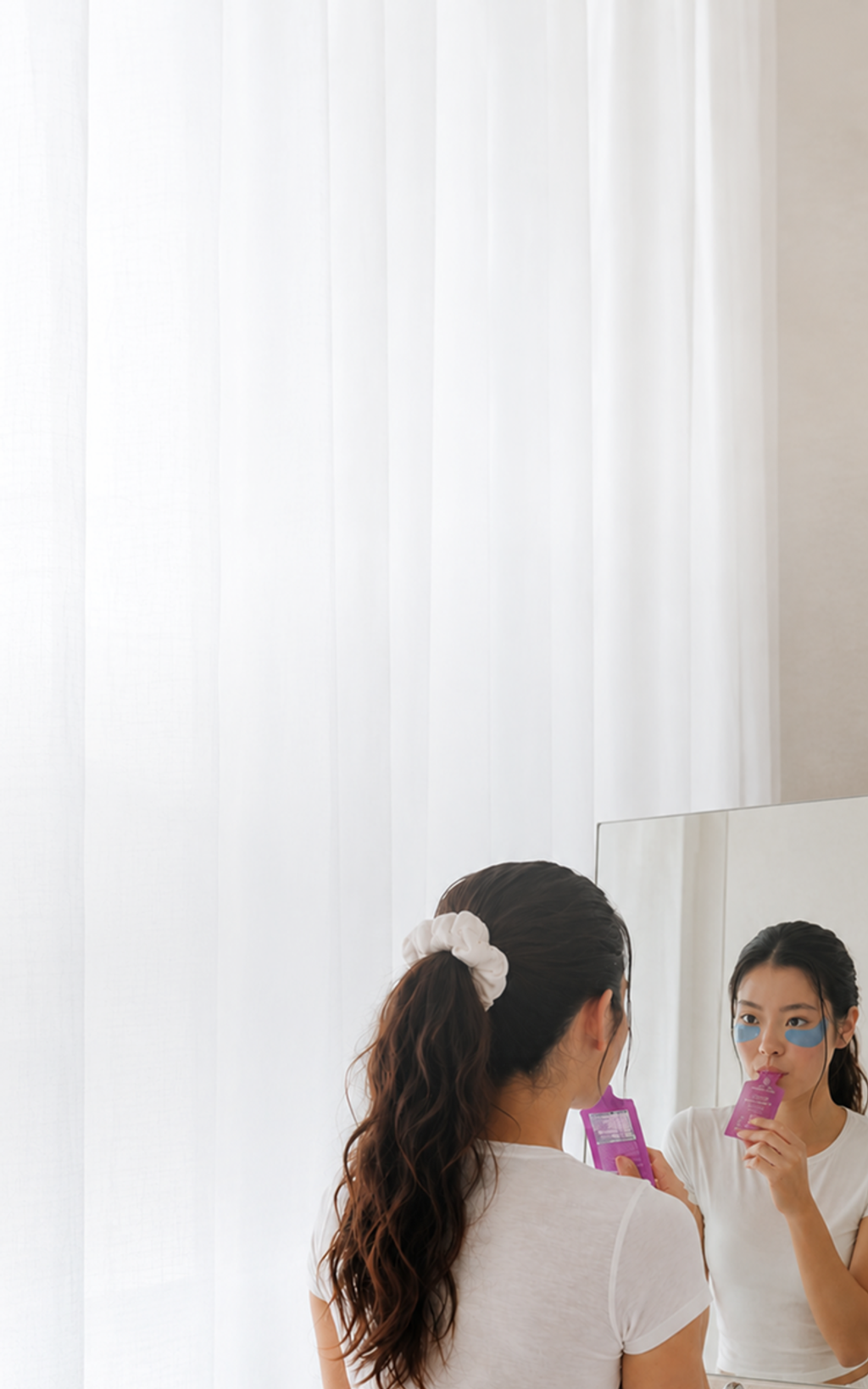 Woman wearing blue under-eye patches holds a pink sachet while looking in a mirror.