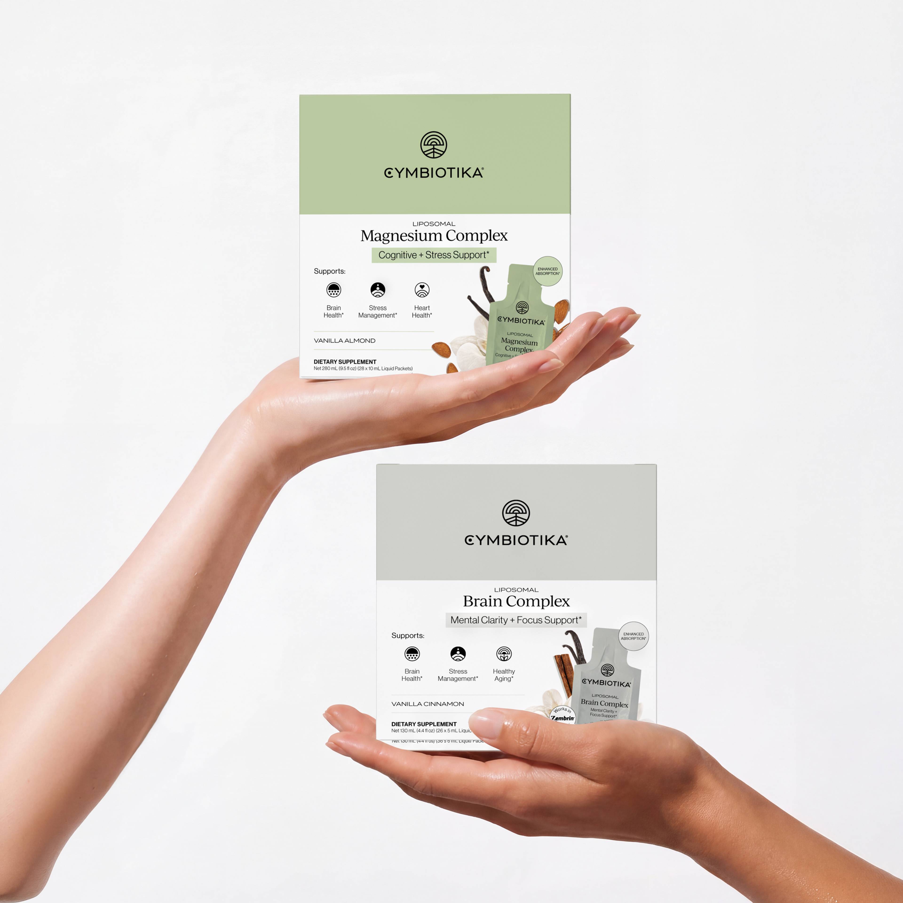 Hands hold Cymbiotika Liposomal Magnesium Complex and Liposomal Brain Complex boxes with matching sachets.