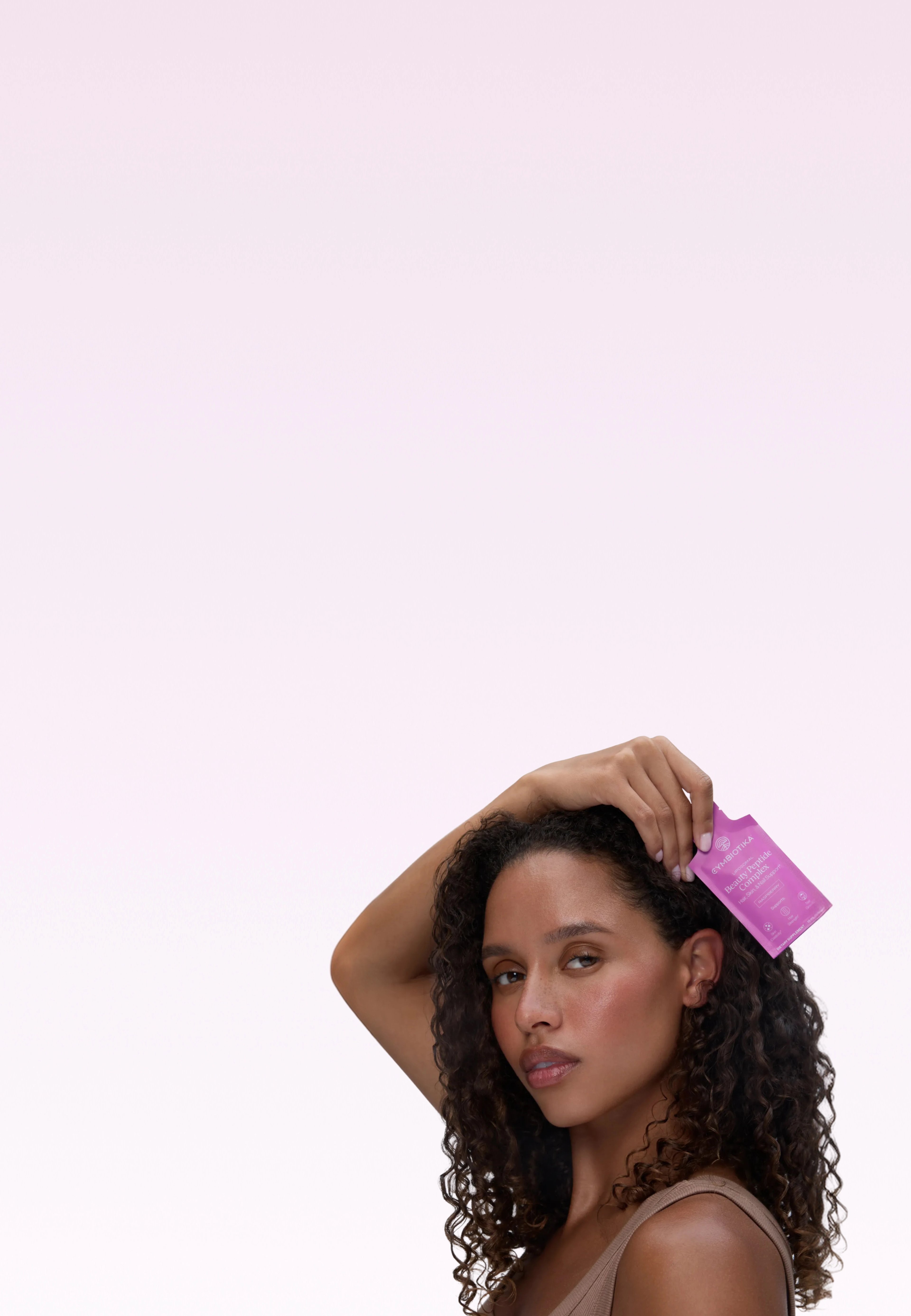 Woman holding a pink Beauty Peptide Complex sachet beside her hair on a pale pink background.