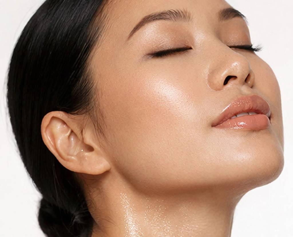 Close-up of a woman with closed eyes and dewy skin against a white background.
