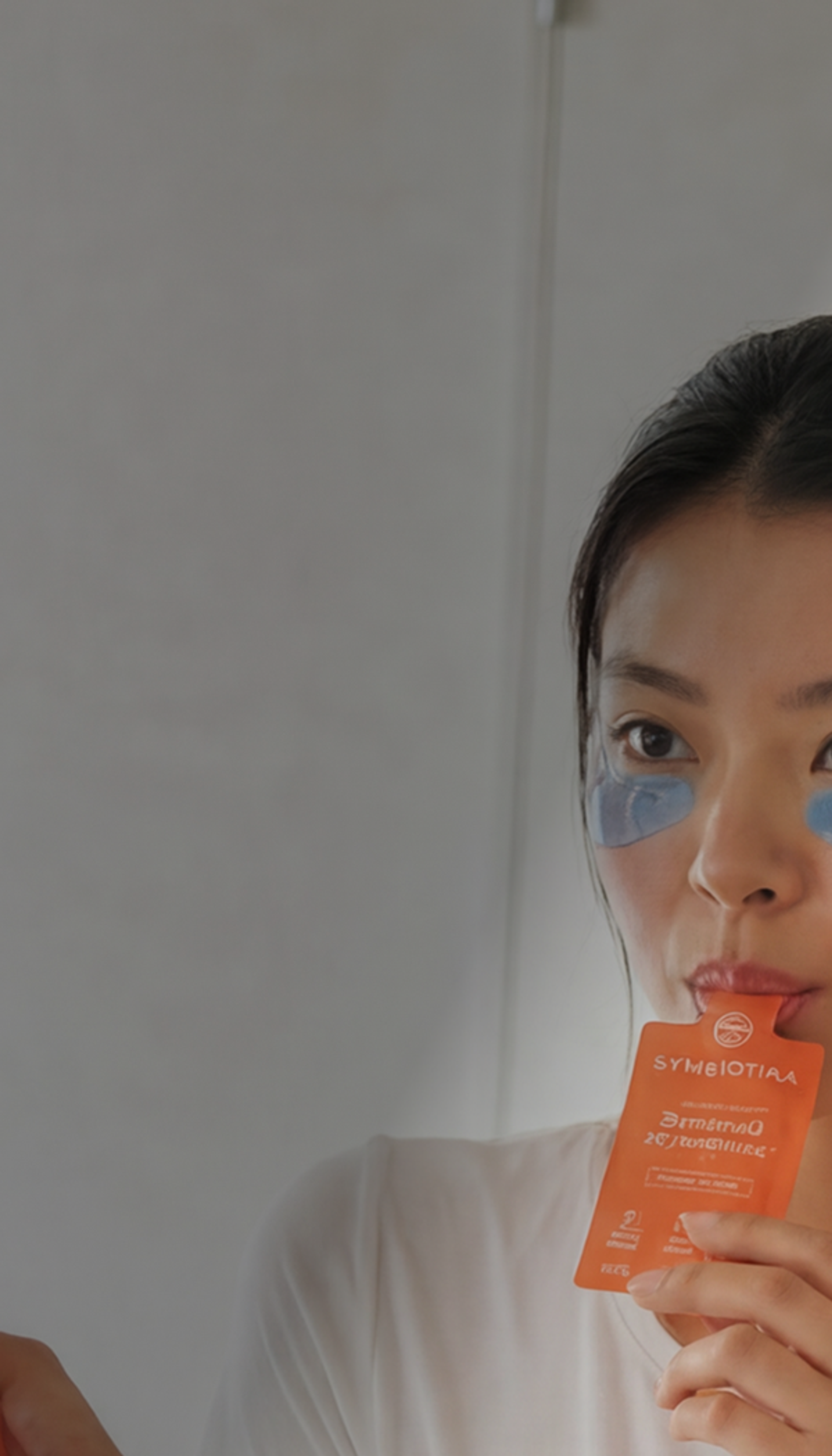 Woman wearing blue under-eye patches holds an orange supplement sachet near her face.