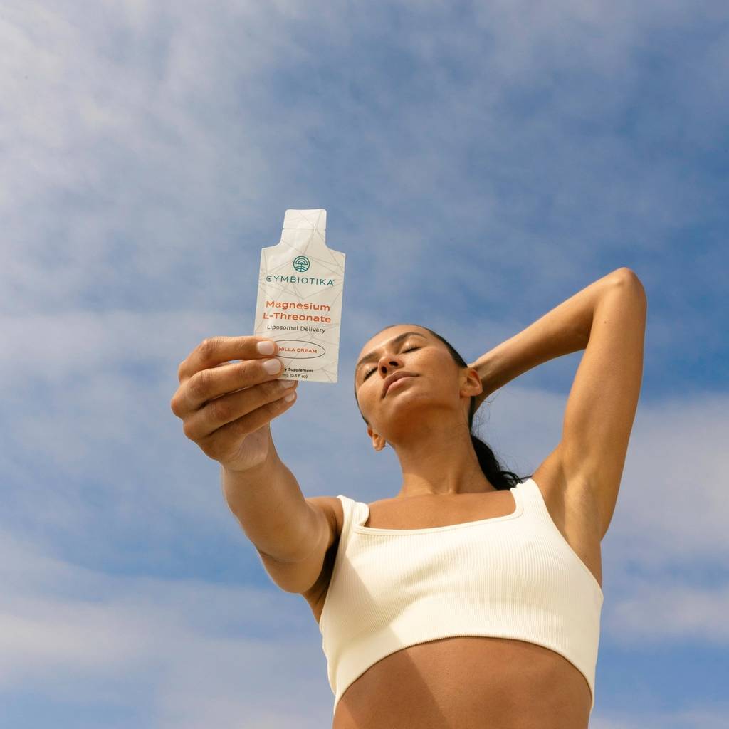 Woman in white activewear holds a Cymbiotika Magnesium L-Threonate pouch against a blue sky.