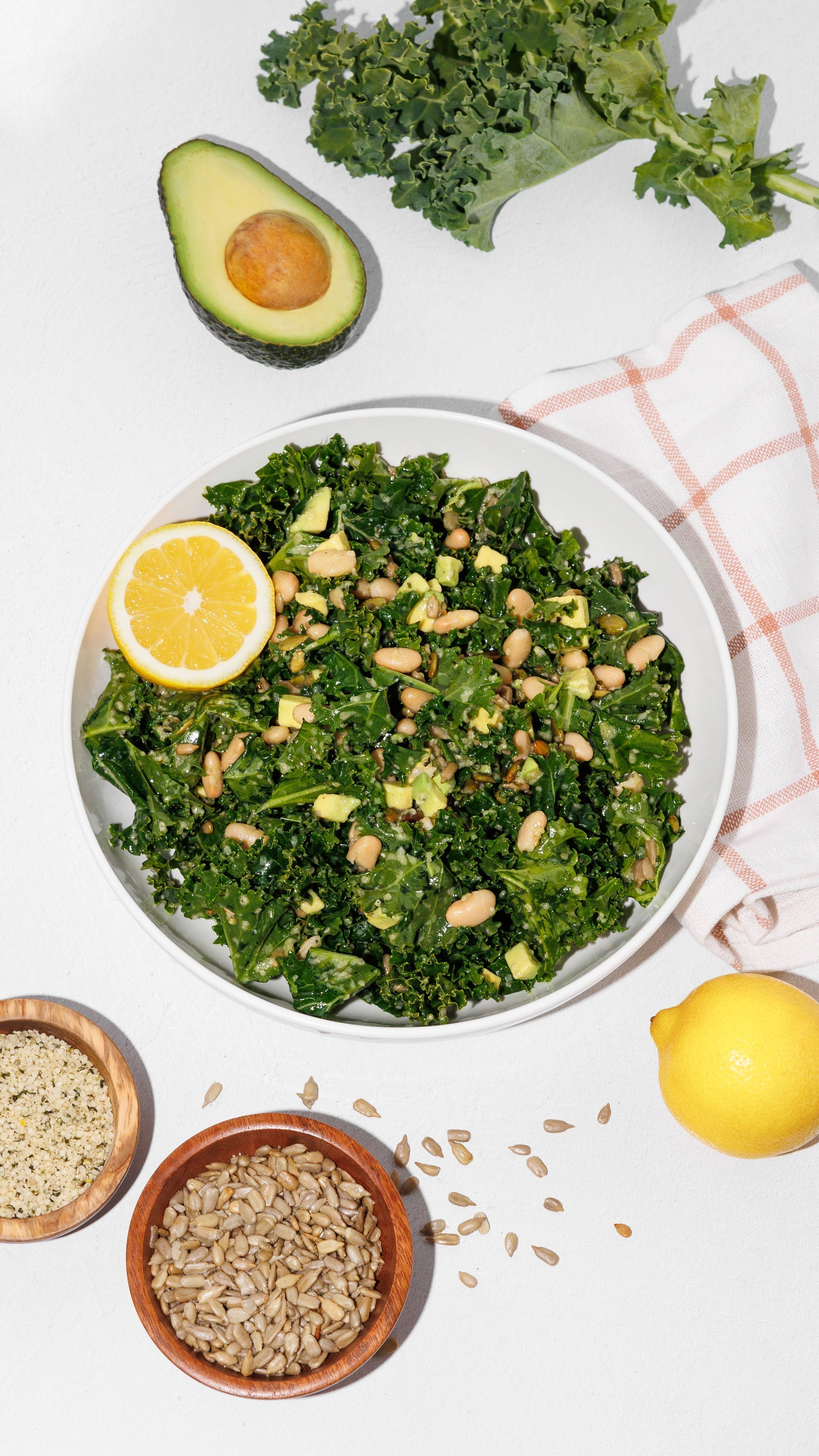 Bowl of kale and white bean salad with avocado, lemon, and sunflower seeds on a white table.