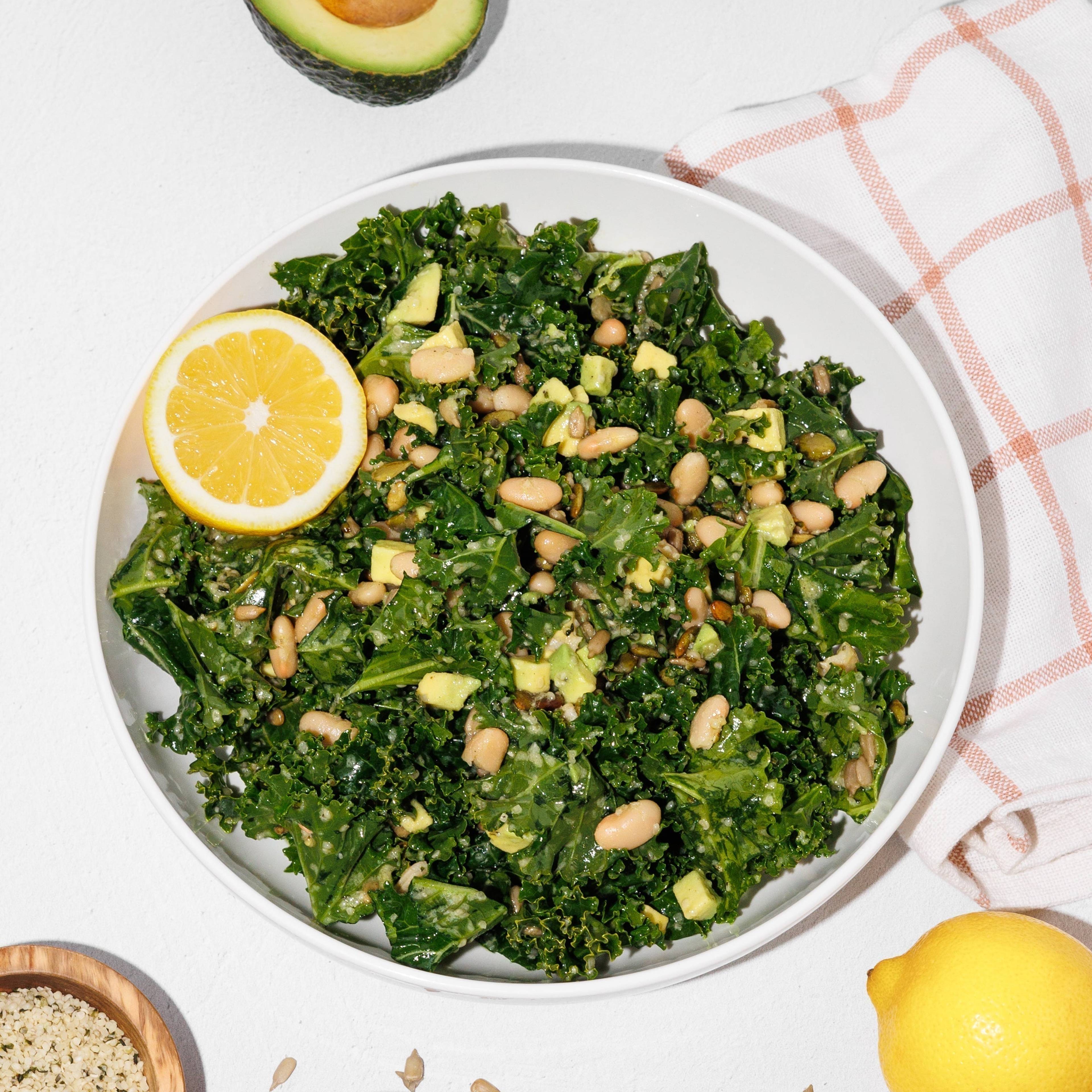 Bowl of kale and white bean salad with avocado, lemon, and sunflower seeds on a white table.