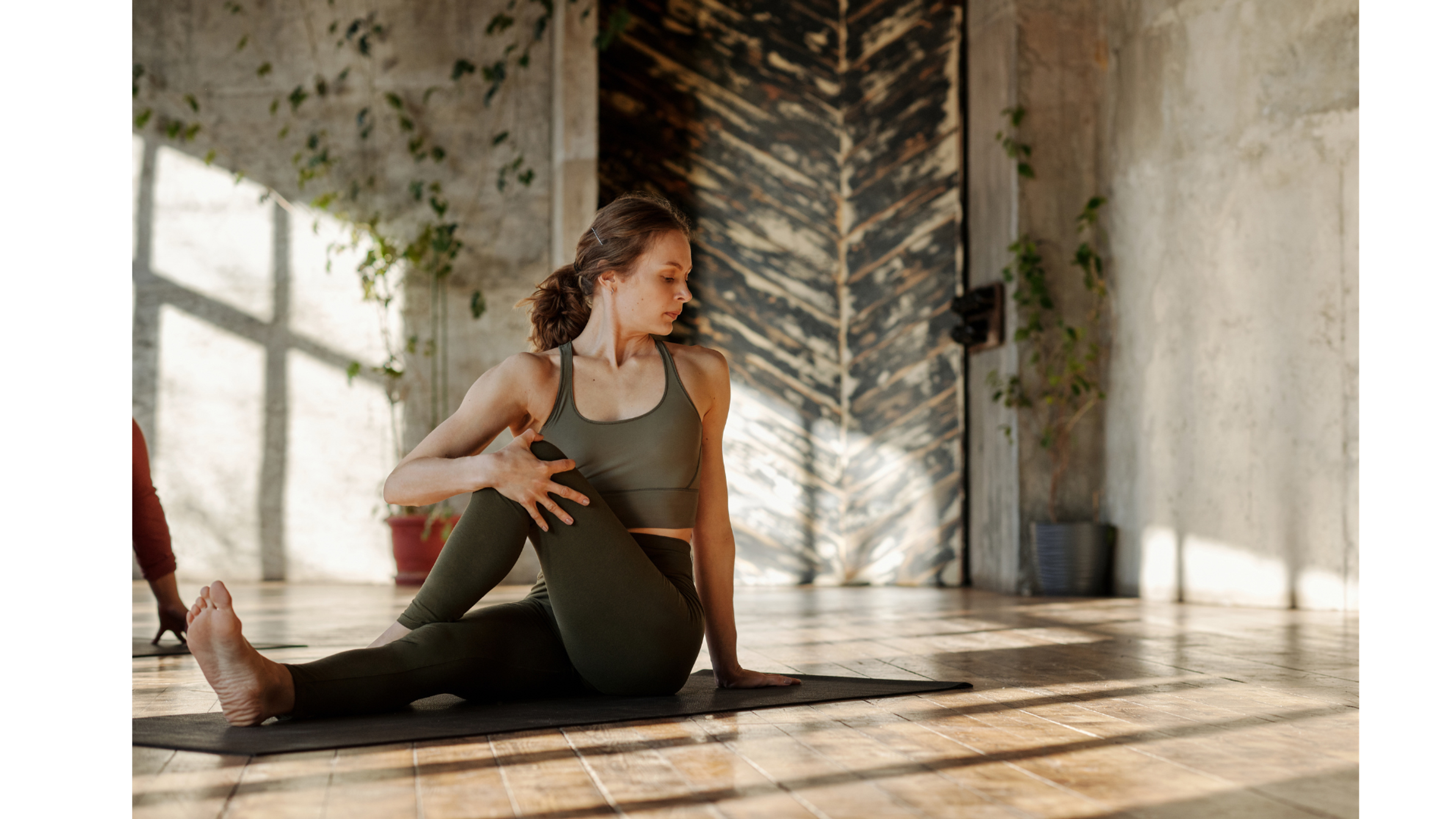 Woman in dark activewear doing a seated spinal twist on a yoga mat in a sunlit studio.