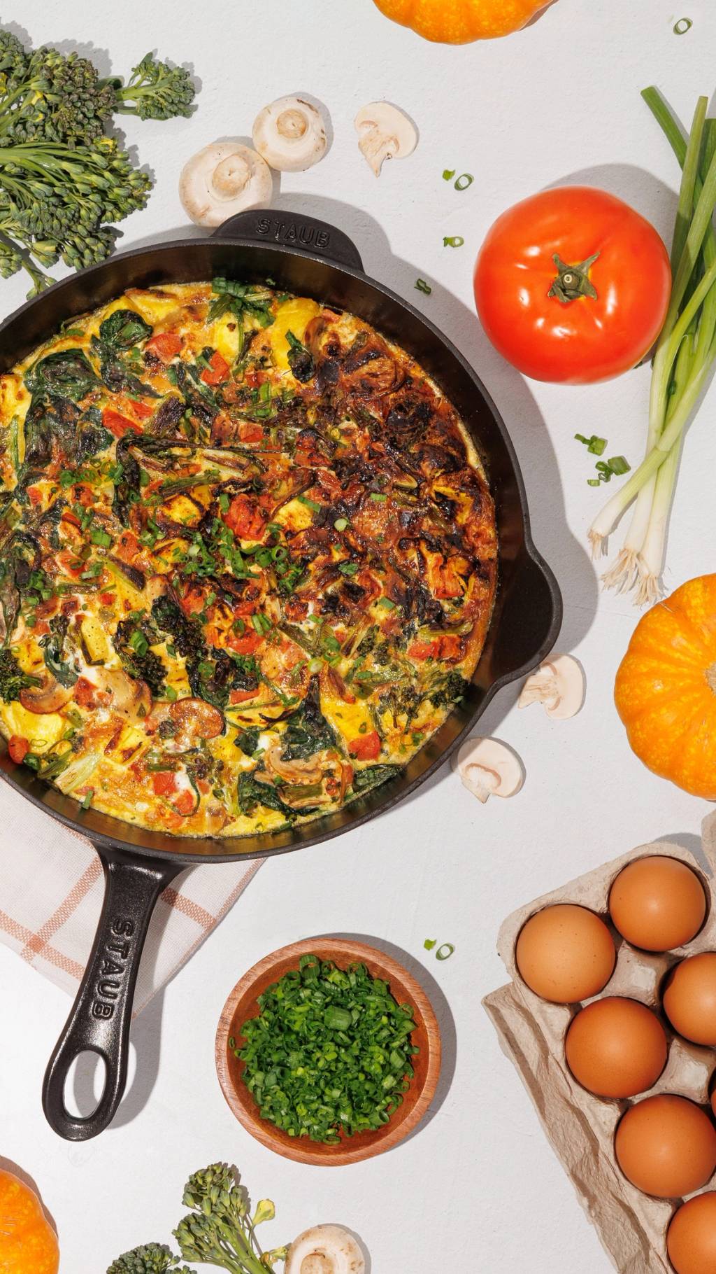 Vegetable frittata in a dark skillet surrounded by broccoli, mushrooms, a tomato, scallions, pumpkins, and eggs.