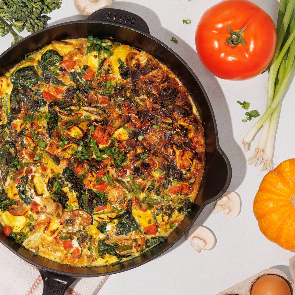 Vegetable frittata in a dark skillet surrounded by broccoli, mushrooms, a tomato, scallions, pumpkins, and eggs.