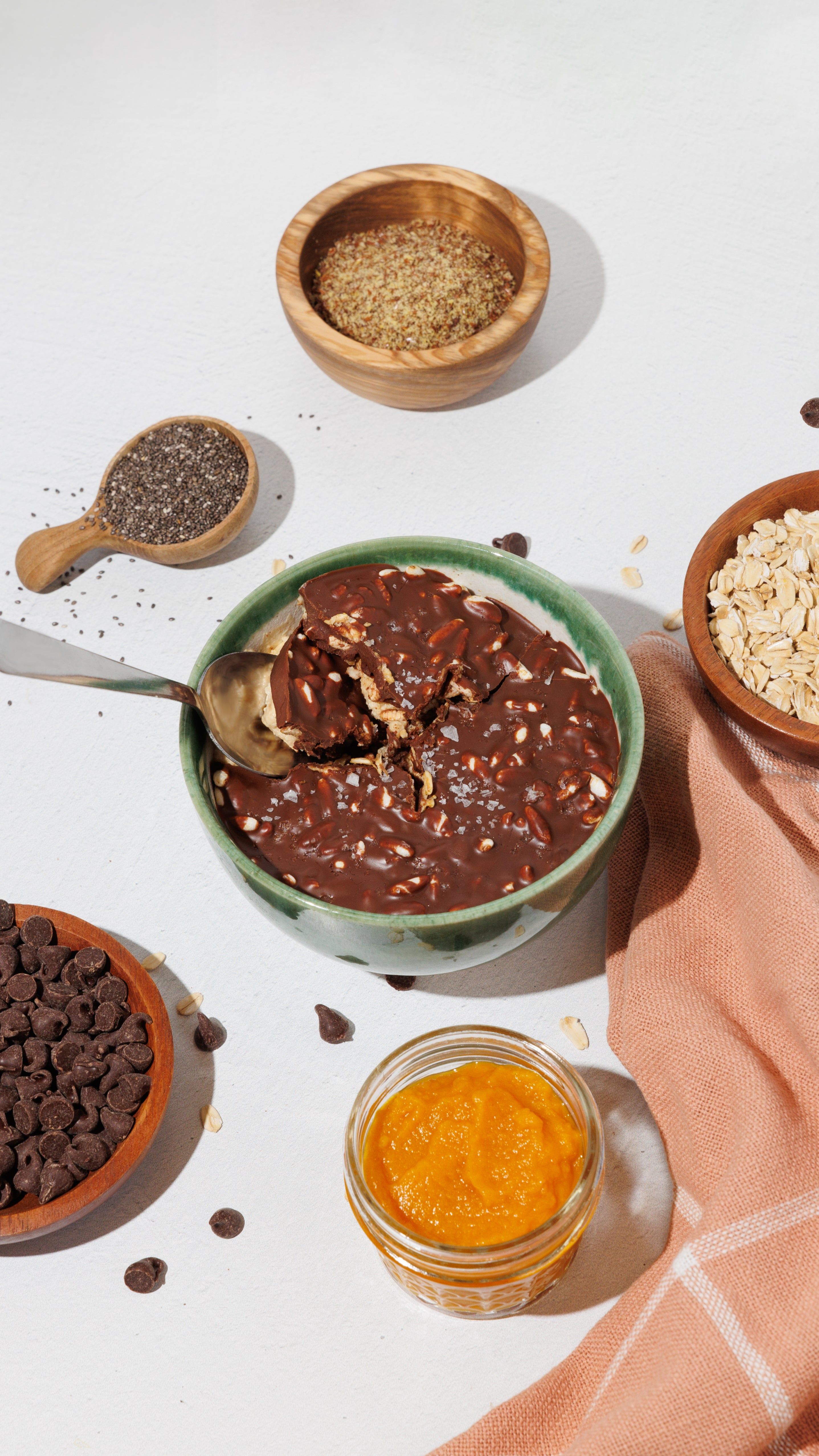 Bowl of chocolate overnight oats with a spoon, surrounded by pumpkin puree, chia seeds, rolled oats, and chocolate chips.