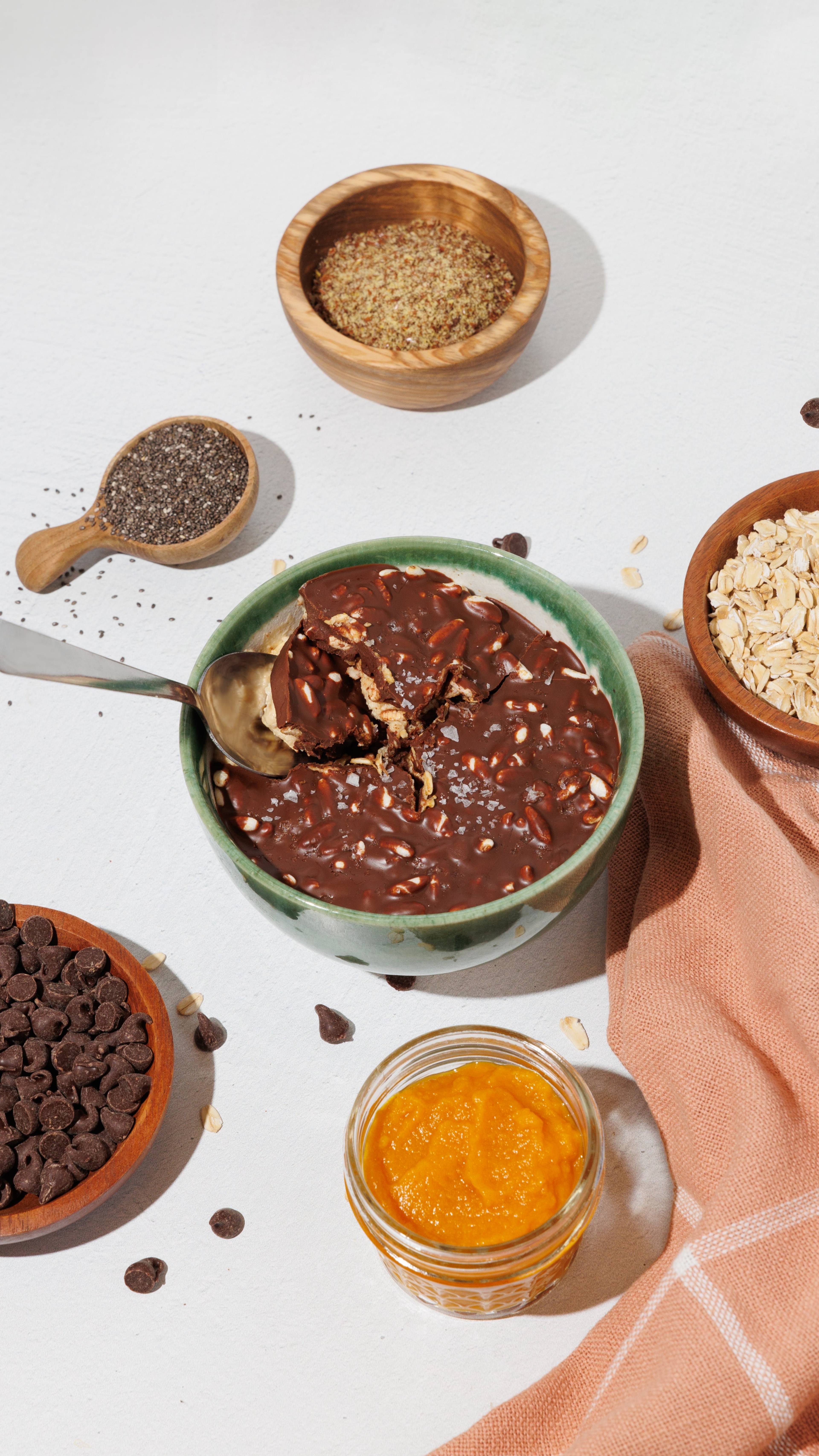Bowl of chocolate overnight oats with a spoon, surrounded by pumpkin puree, chia seeds, rolled oats, and chocolate chips.