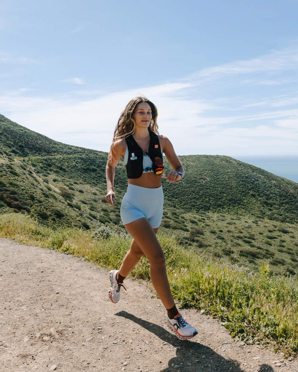 Woman jogging on a sunlit coastal trail, wearing a black hydration vest and light blue athletic shorts.