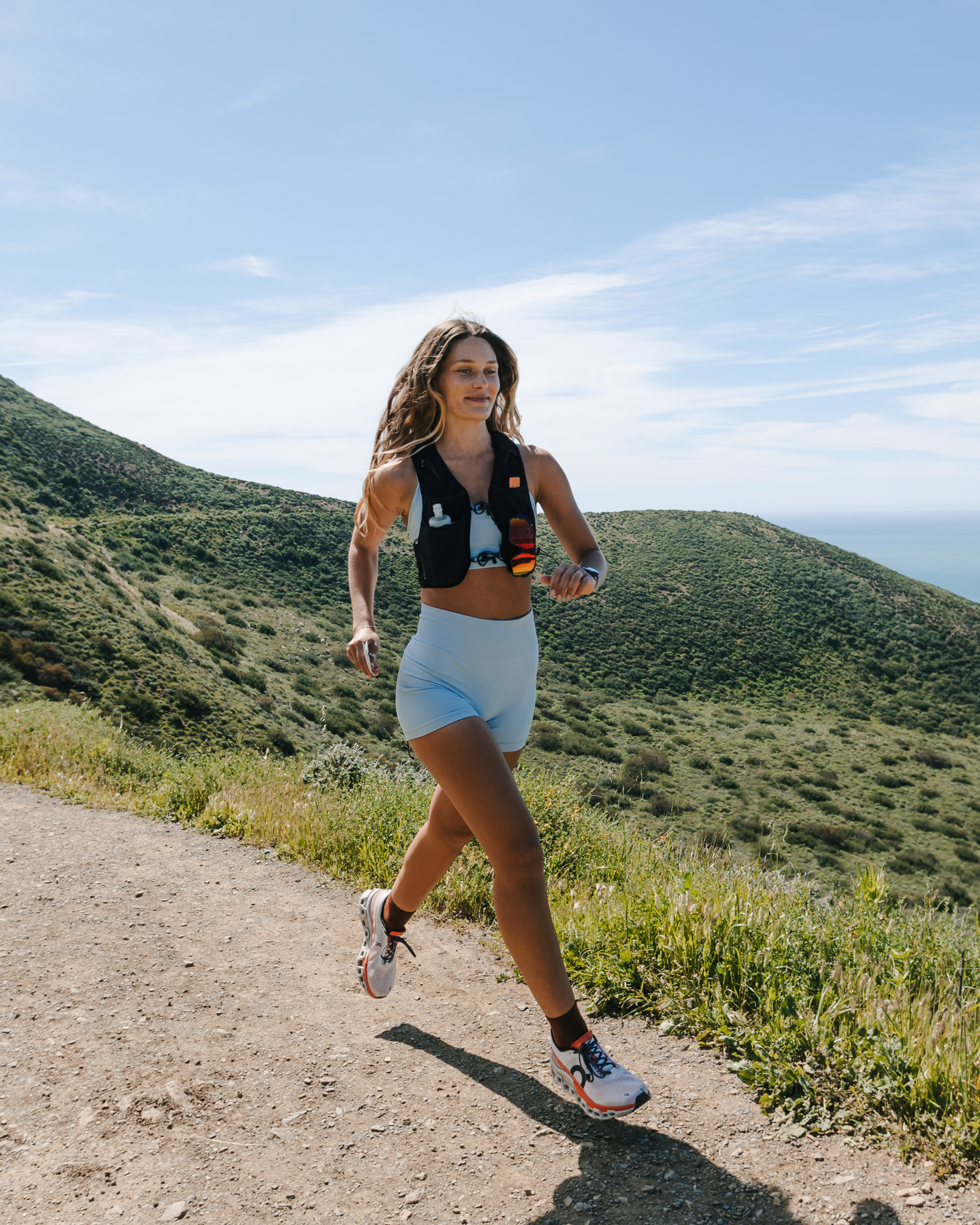 Woman jogging on a sunlit coastal trail, wearing a black hydration vest and light blue athletic shorts.