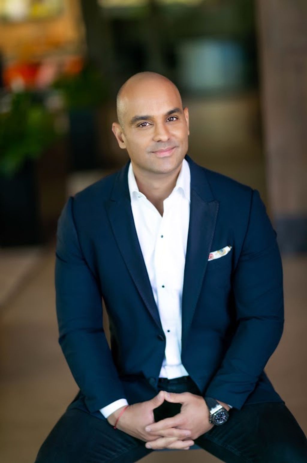 Bald man in a navy suit and white shirt seated indoors with hands clasped.