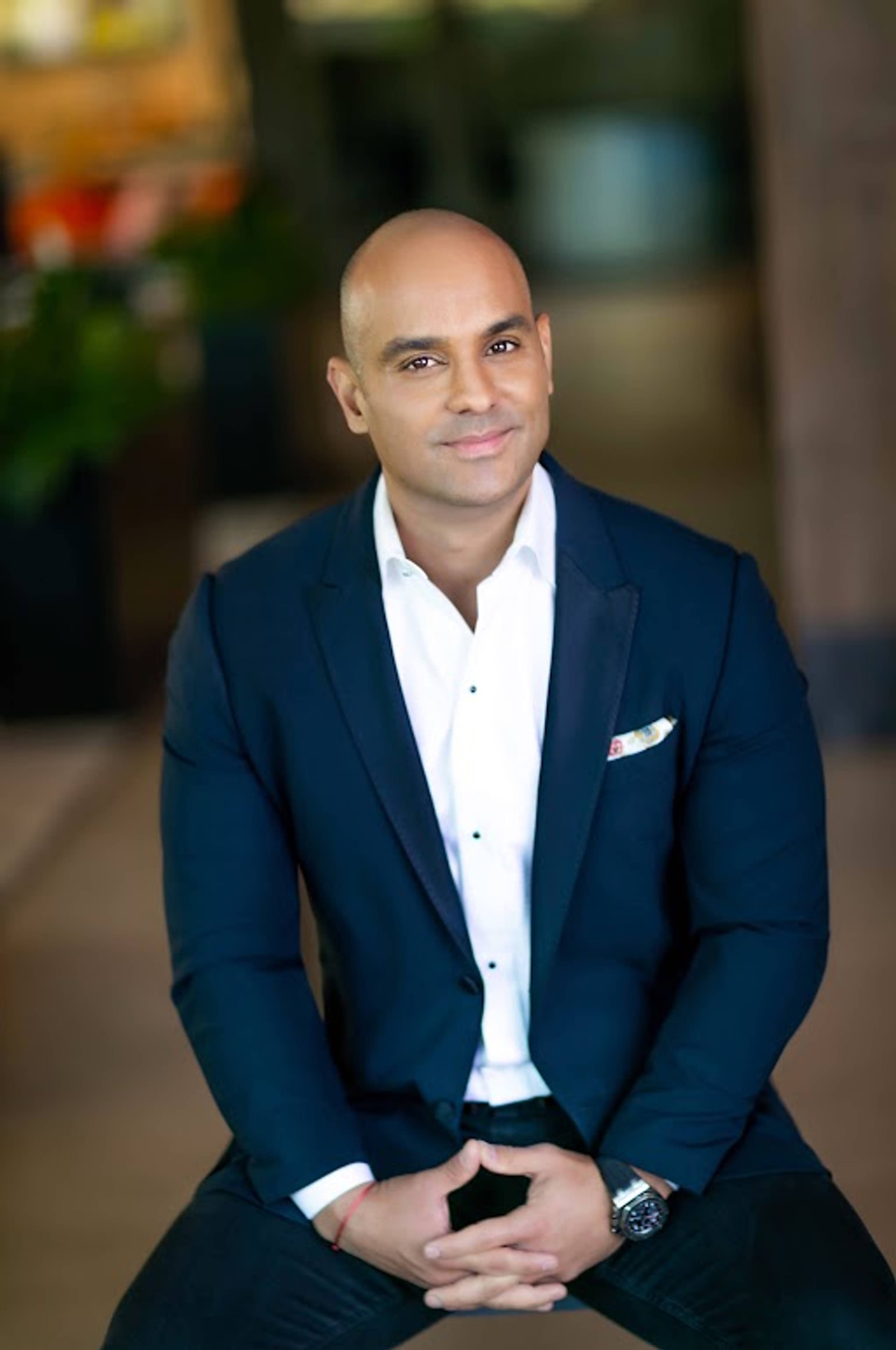 Bald man in a navy suit and white shirt seated indoors with hands clasped.