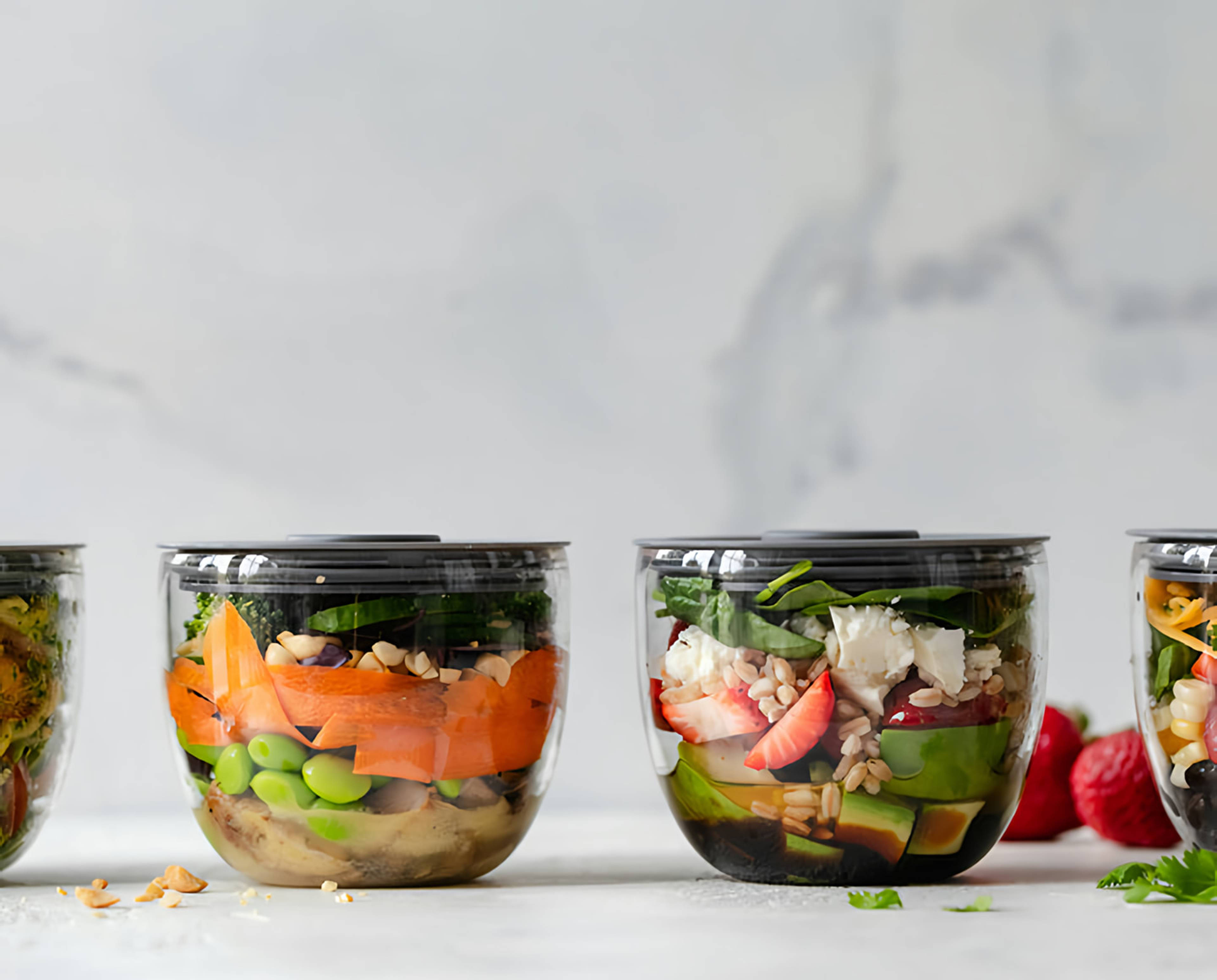 Clear containers of layered salads with greens, avocado, strawberries, grains, and vegetables.