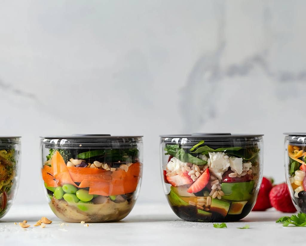 Clear containers of layered salads with greens, avocado, strawberries, grains, and vegetables.