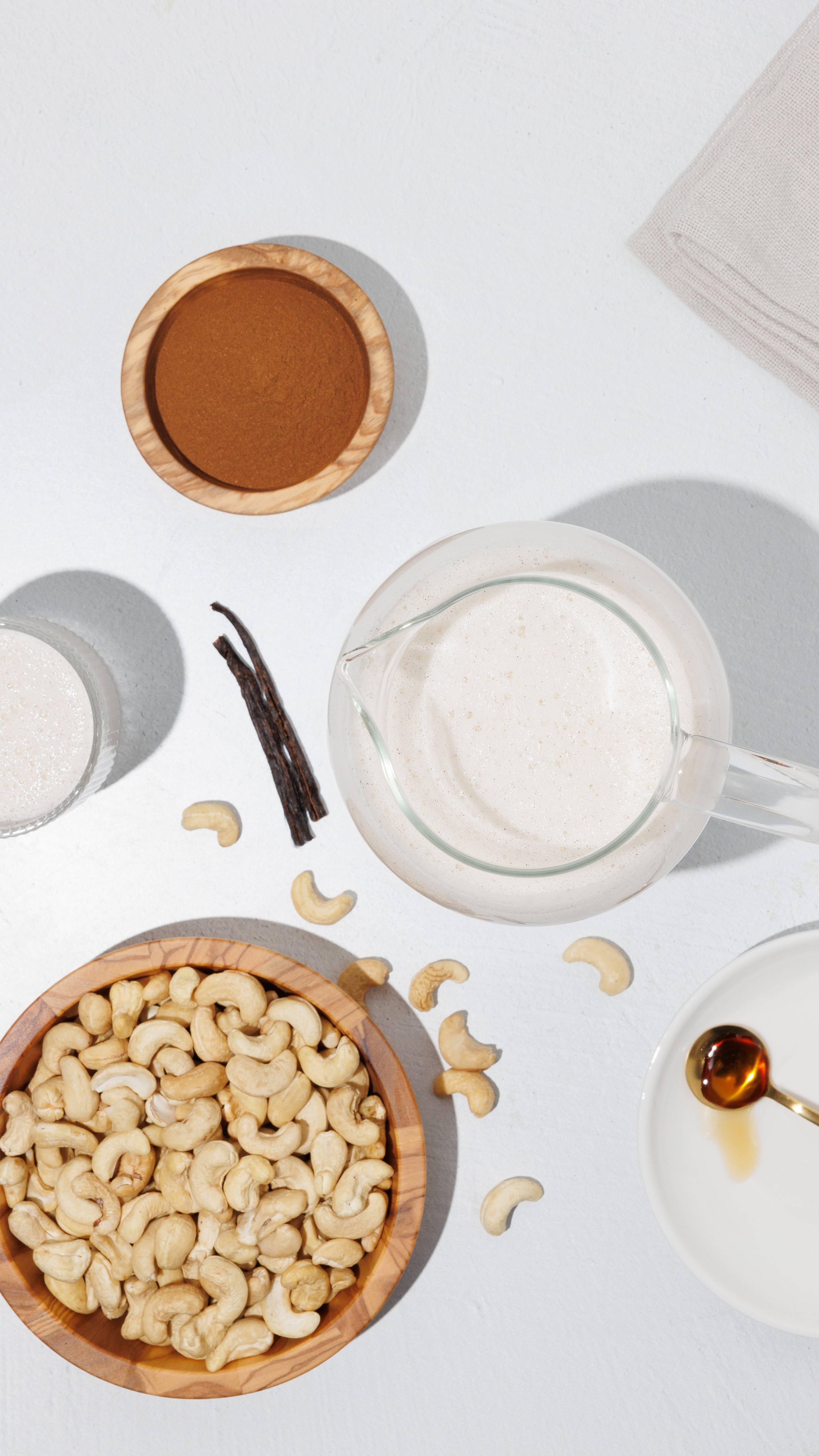 Overhead arrangement of cashews, dark vanilla pods, a bowl of brown powder, and a pitcher of pale, creamy-looking liquid.