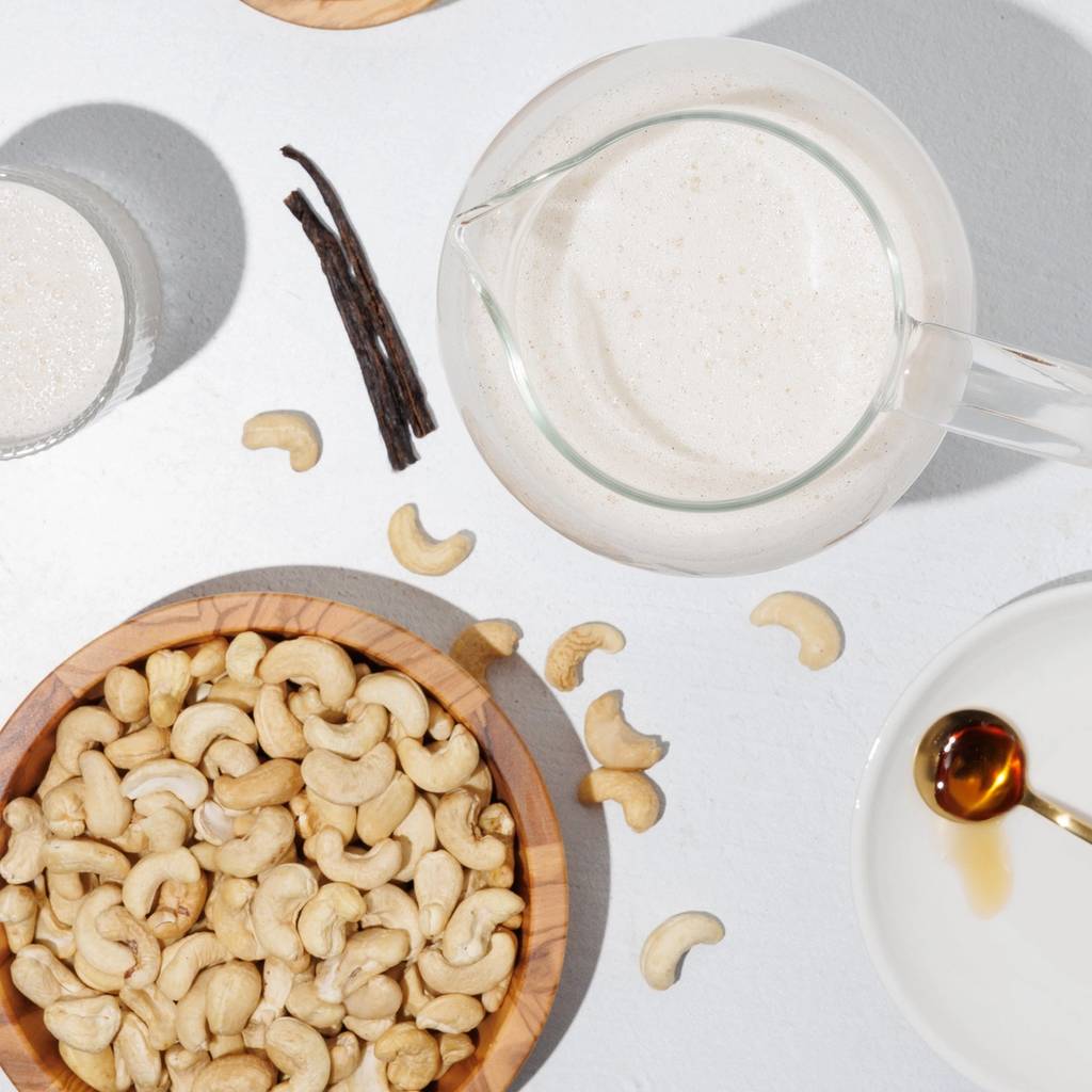 Overhead arrangement of cashews, dark vanilla pods, a bowl of brown powder, and a pitcher of pale, creamy-looking liquid.
