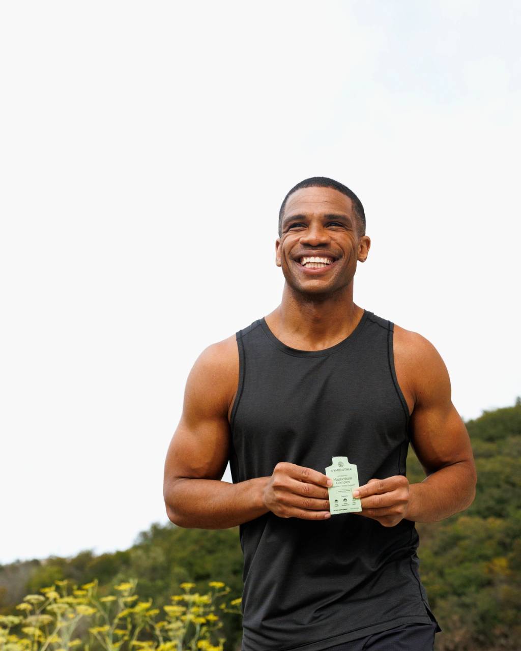 Smiling man in a black tank top holds a small supplement packet outdoors.