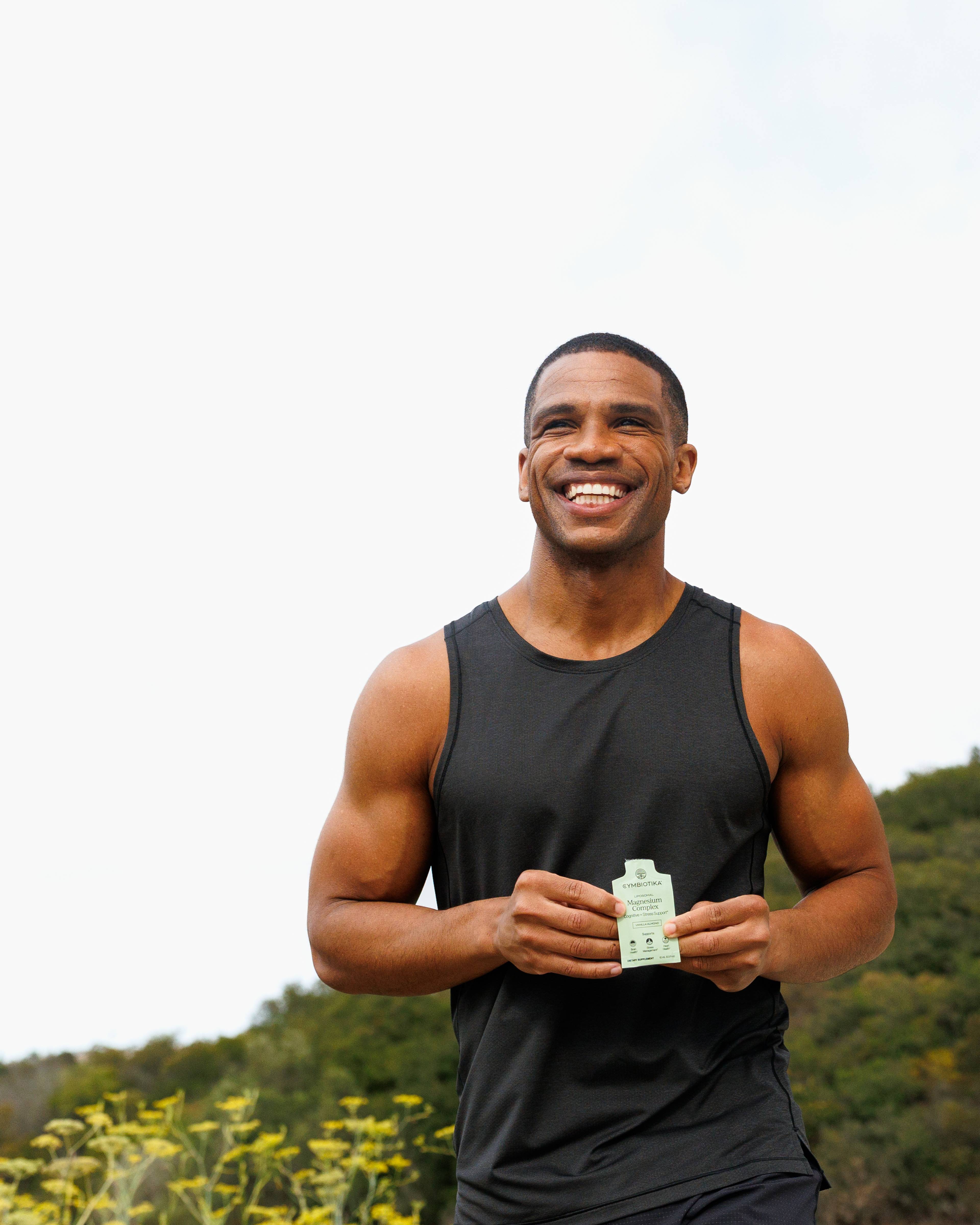 Smiling man in a black tank top holds a small supplement packet outdoors.