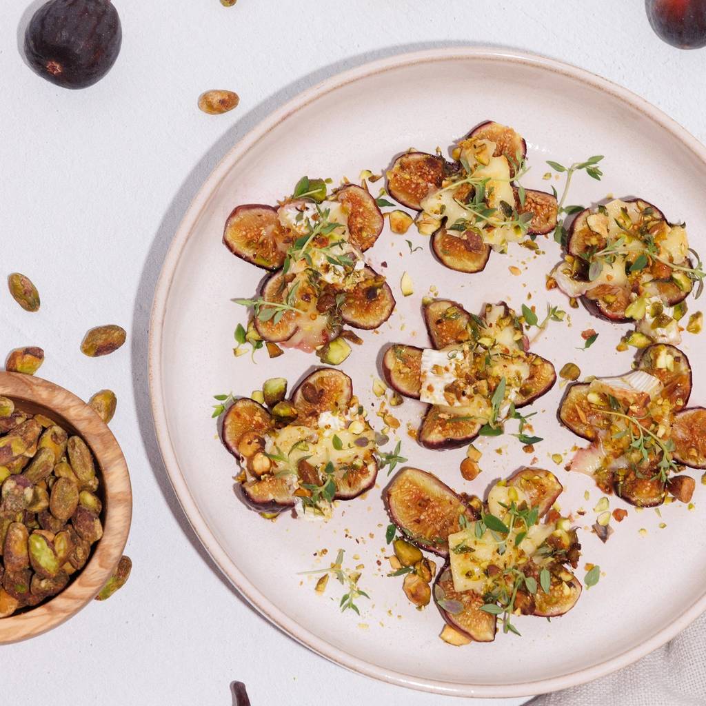 Plate of baked fig and brie bites topped with pistachios and herbs, surrounded by fresh figs.
