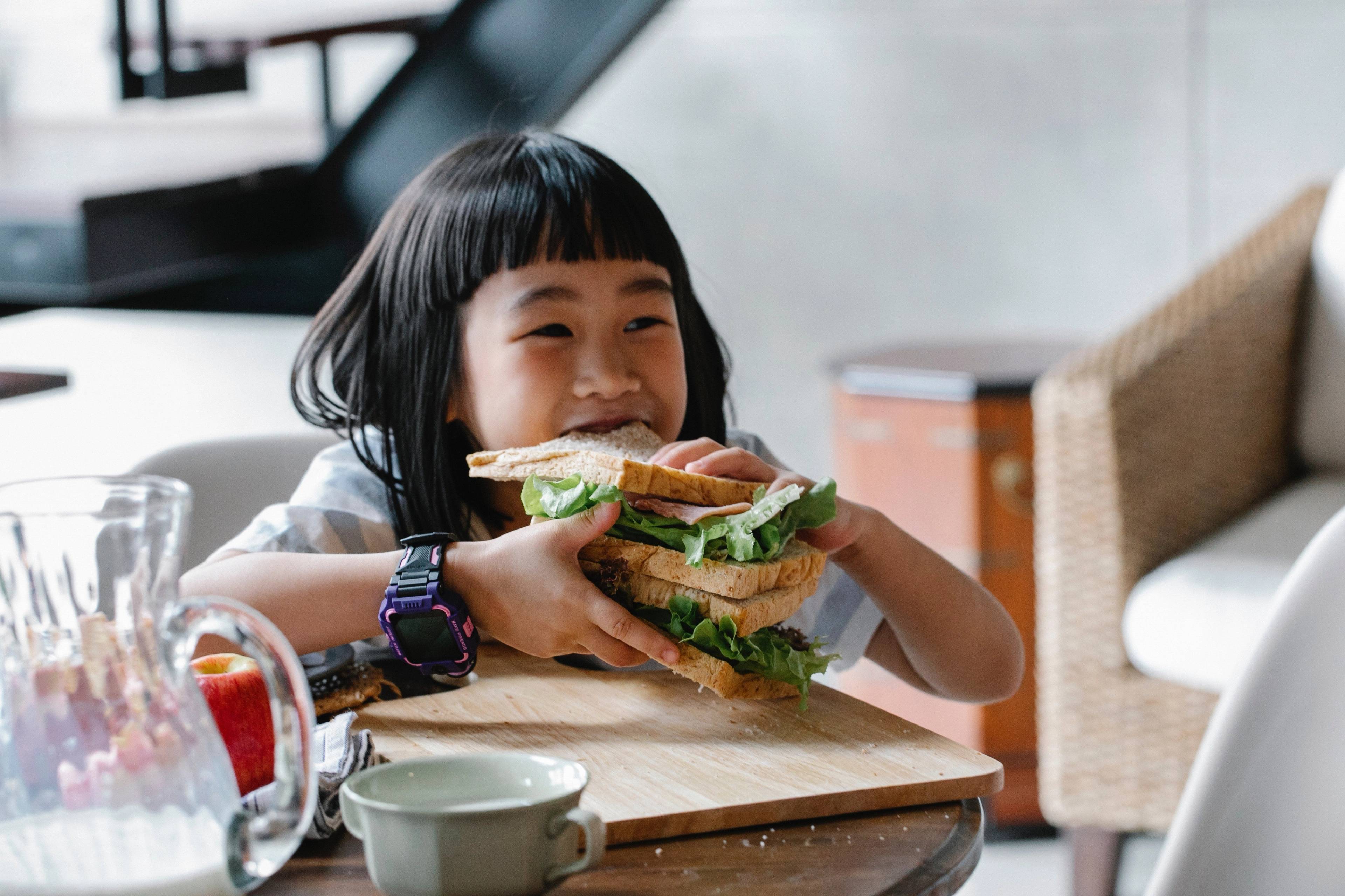 Smiling child holding a large sandwich with leafy greens at a dining table
