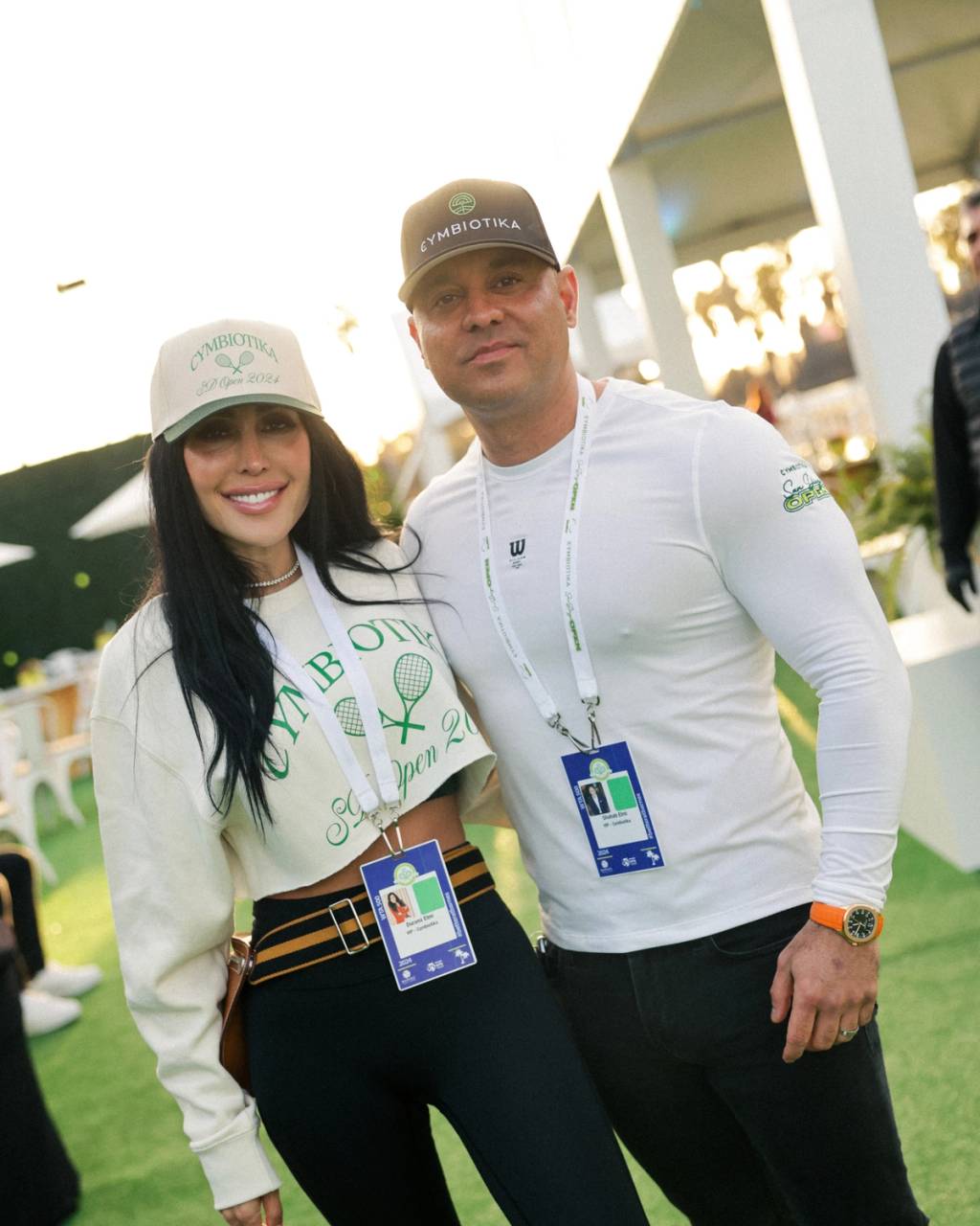 Two attendees wearing Cymbiotika hats and event lanyards pose outdoors at a tennis event.