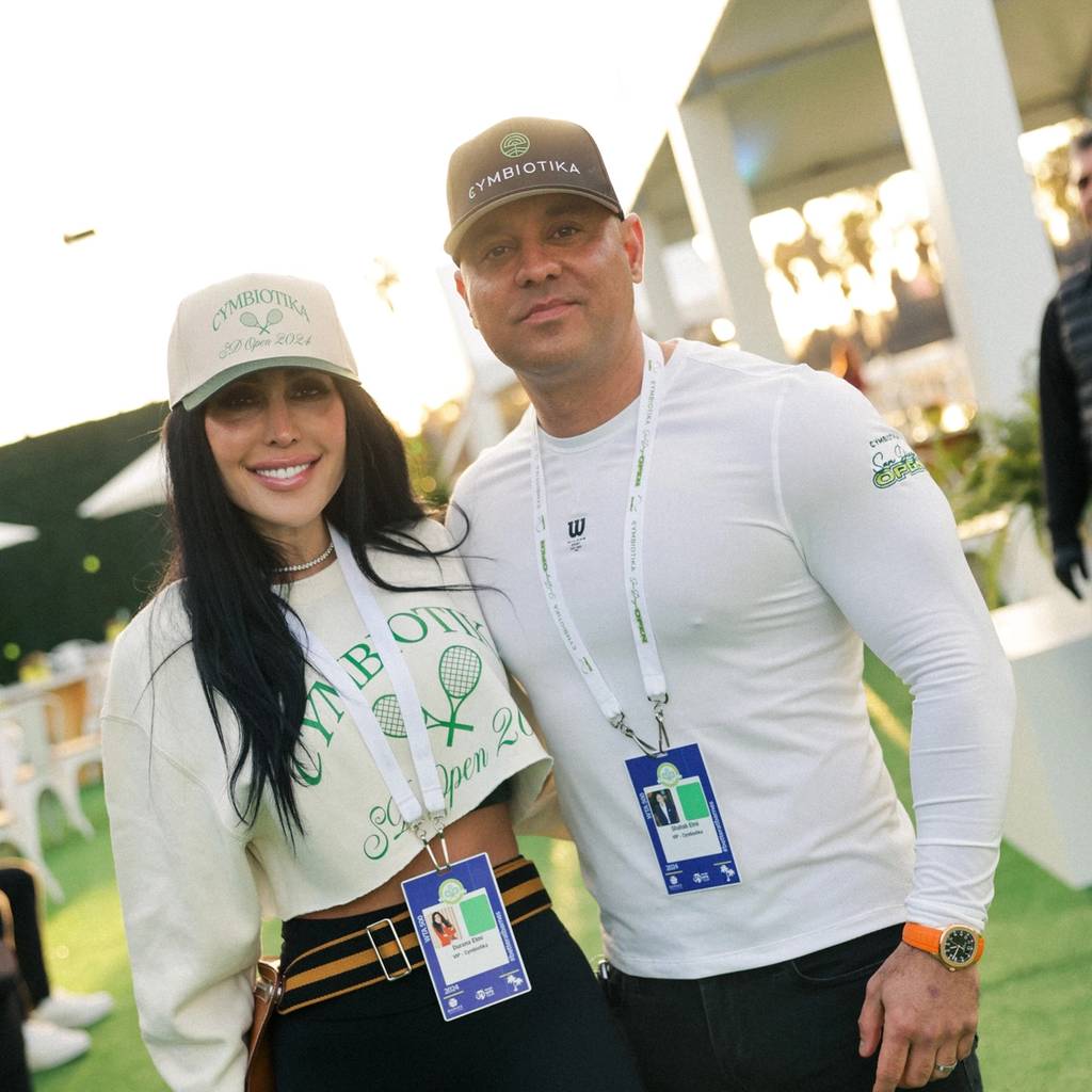Two attendees wearing Cymbiotika hats and event lanyards pose outdoors at a tennis event.