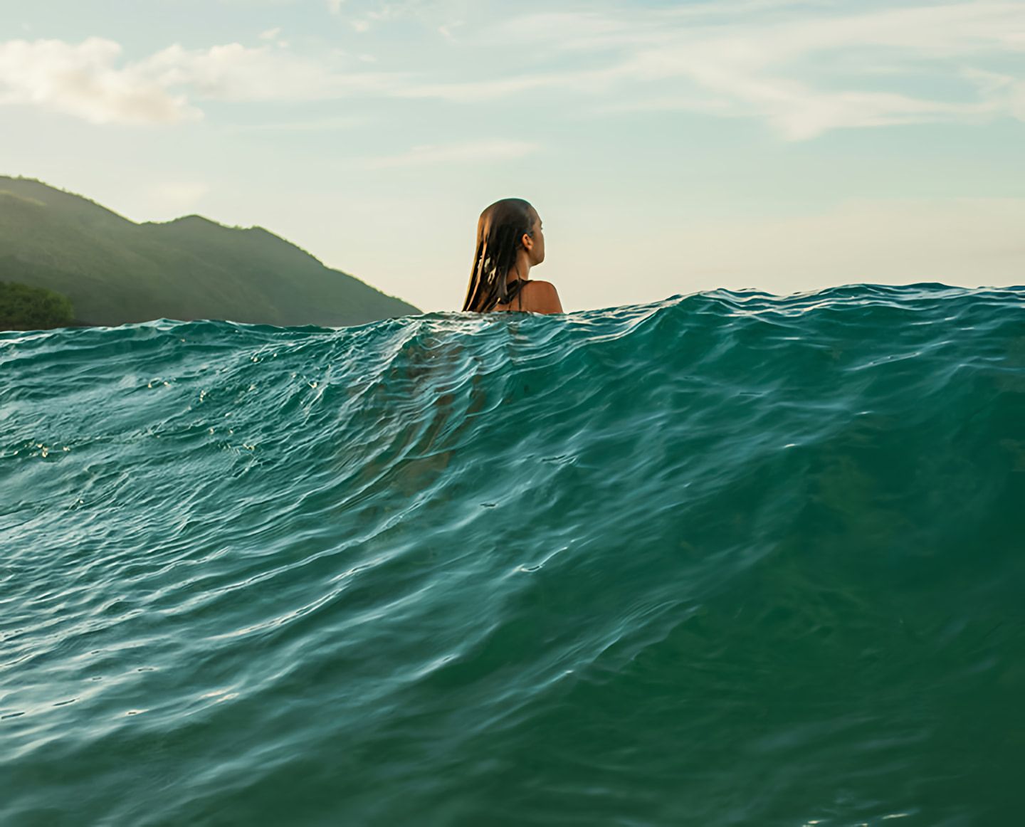 Woman with wet hair swimming in ocean waves near a mountainous shoreline