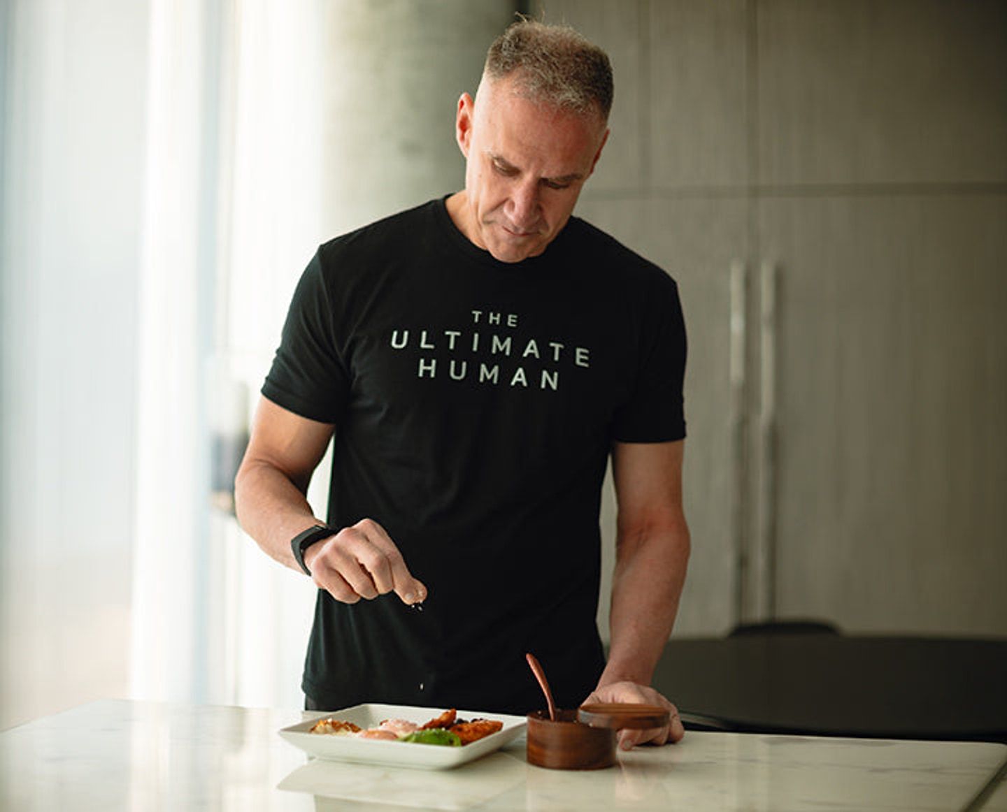 Man in a black T-shirt seasoning food on a plate at a kitchen counter.