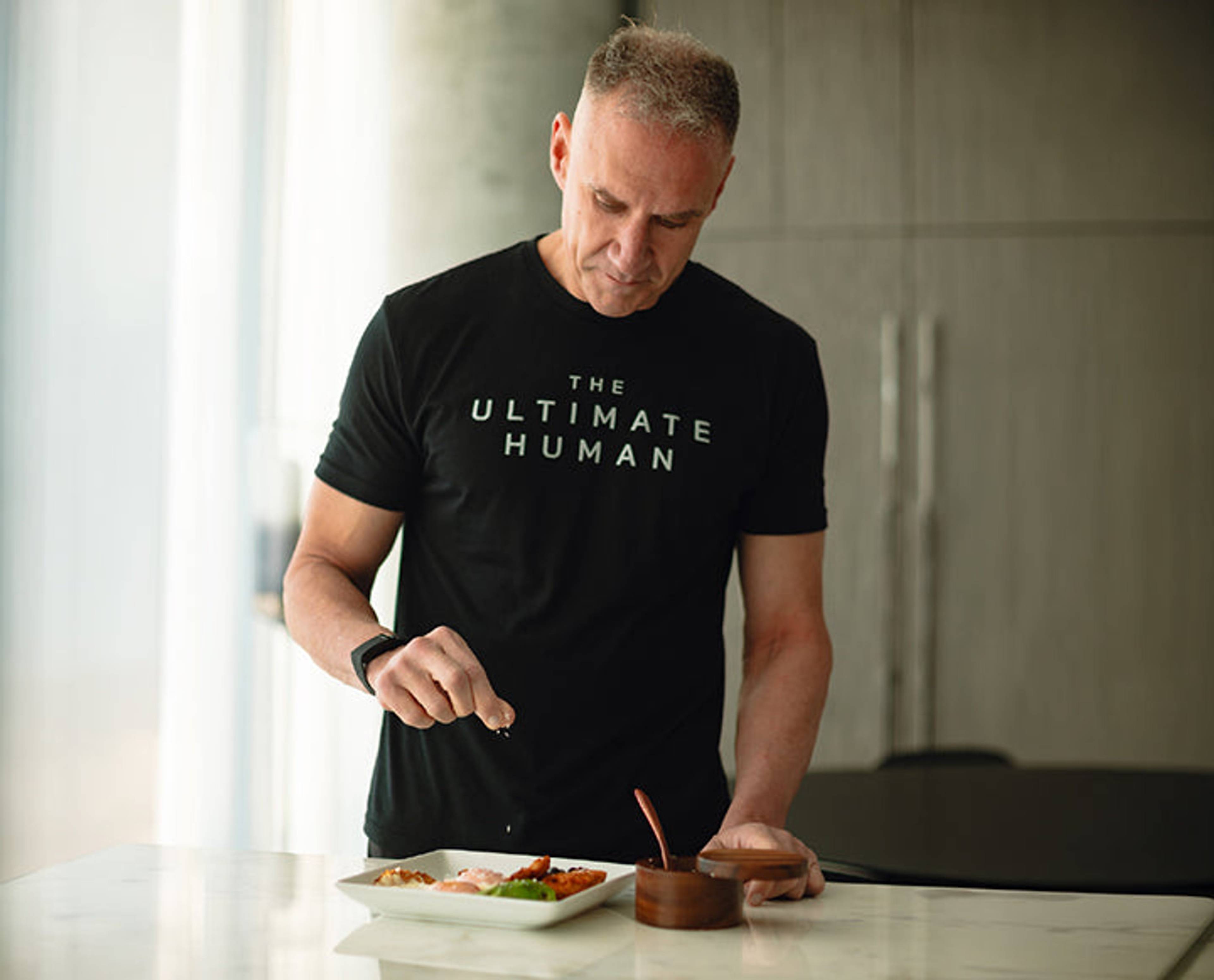 Man in a black T-shirt seasoning food on a plate at a kitchen counter.