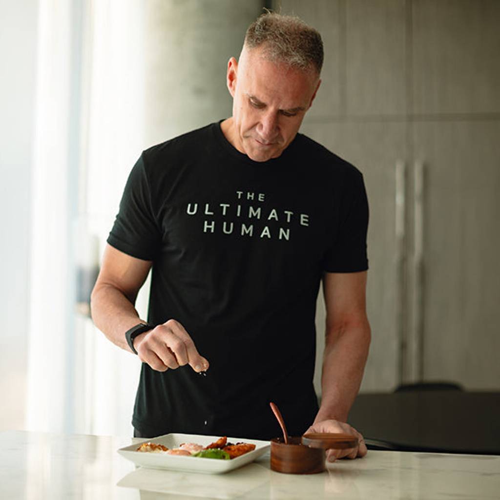 Man in a black T-shirt seasoning food on a plate at a kitchen counter.