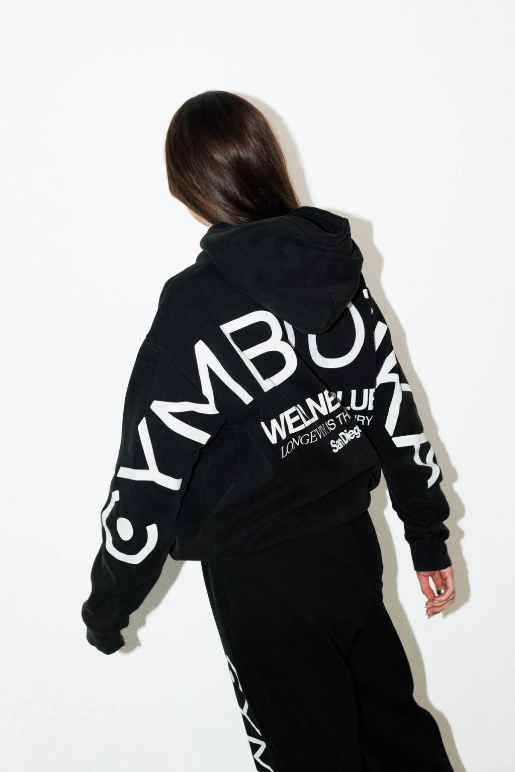 Model shown from behind wearing a black oversized Cymbiotika hoodie with the logo printed in large white letters across the back and sleeves, paired with matching black sweatpants.