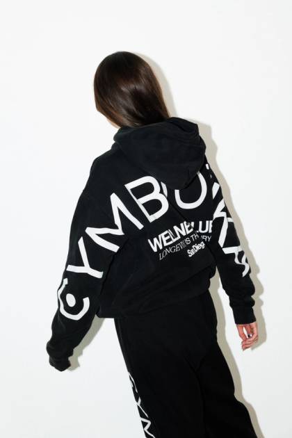 Model shown from behind wearing a black oversized Cymbiotika hoodie with the logo printed in large white letters across the back and sleeves, paired with matching black sweatpants.