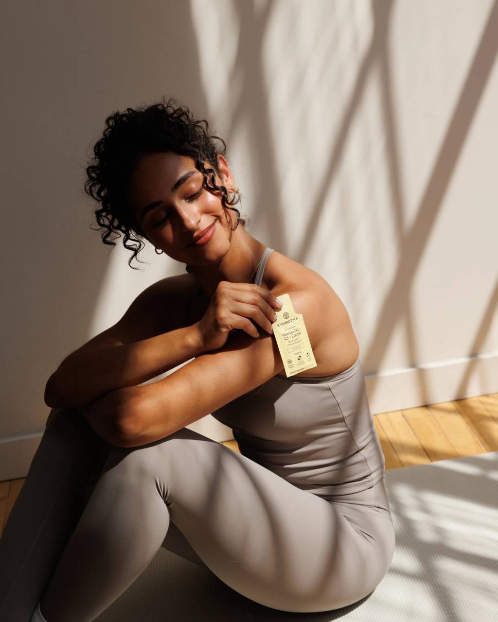 Woman in gray activewear sits on a yoga mat holding a Cymbiotika Liposomal Vitamin D3 + K2 + CoQ10 packet.
