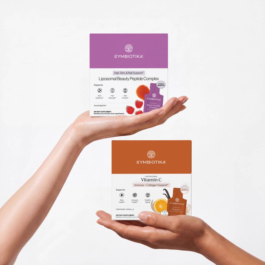 Hands hold Cymbiotika Liposomal Beauty Peptide Complex and Liposomal Vitamin C boxes against a white background.