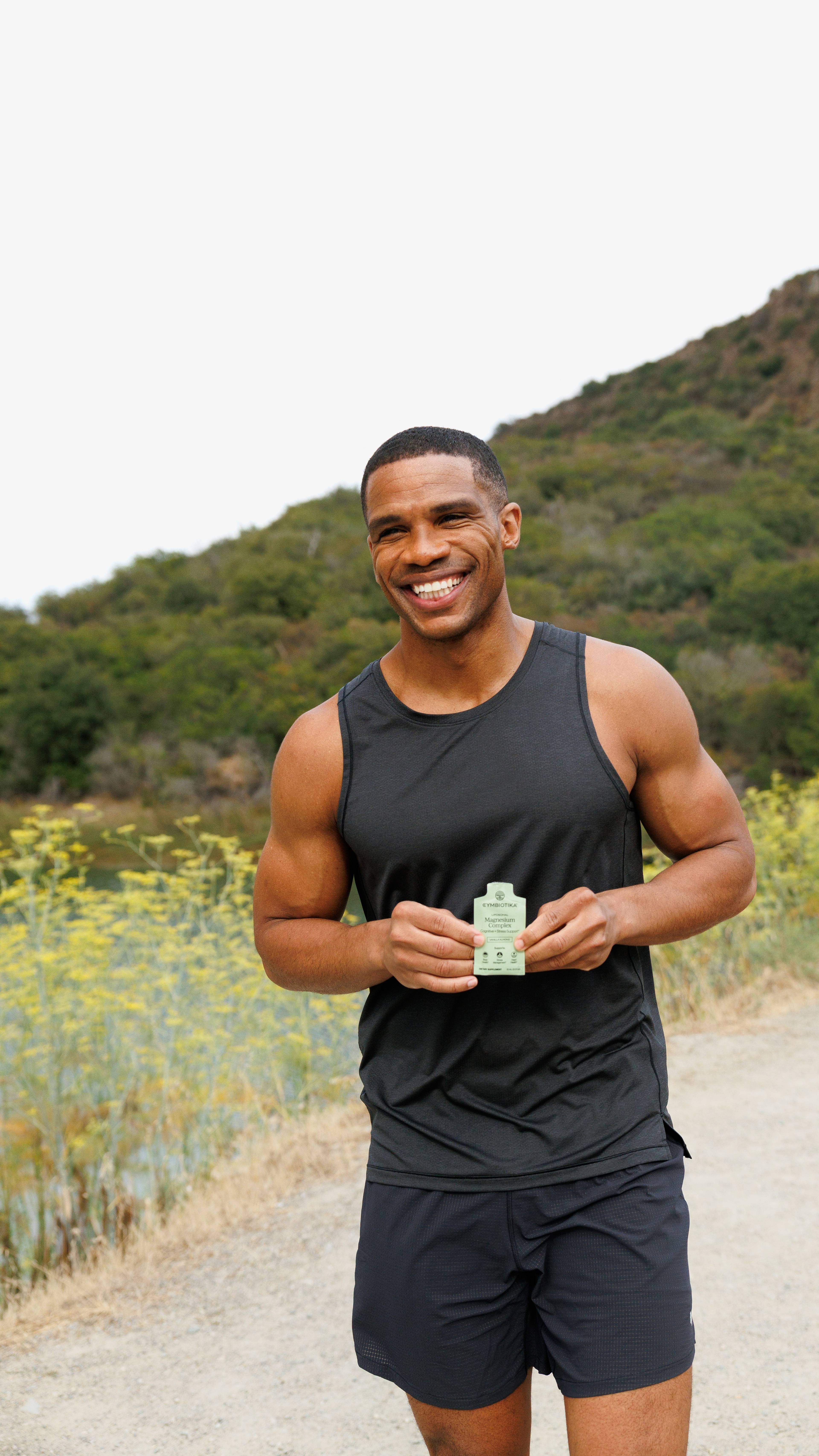 Smiling man in athletic clothing holds a small green supplement packet on a trail.