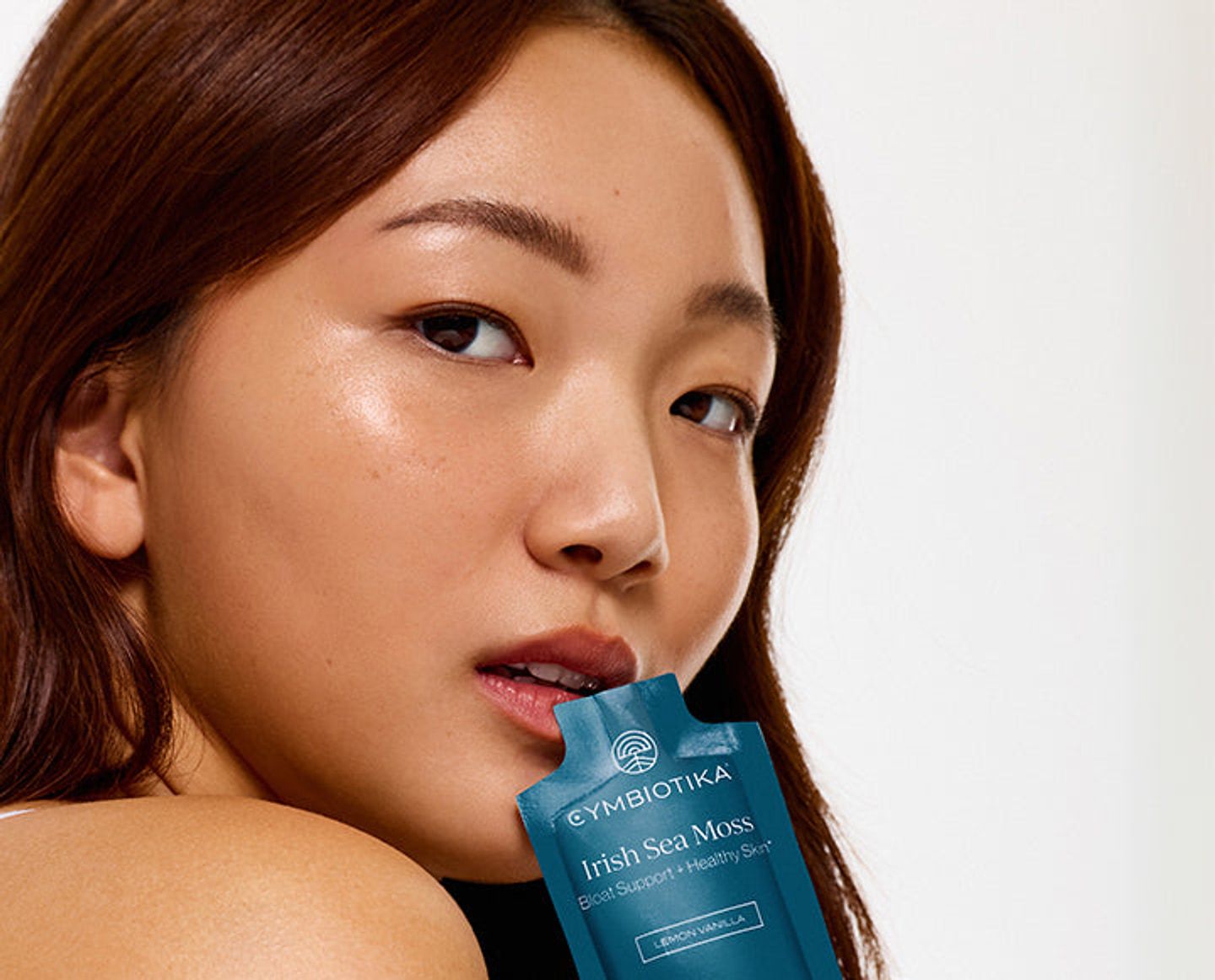 A woman poses with a blue Cymbiotika Irish Sea Moss sachet near her lips against a light background.