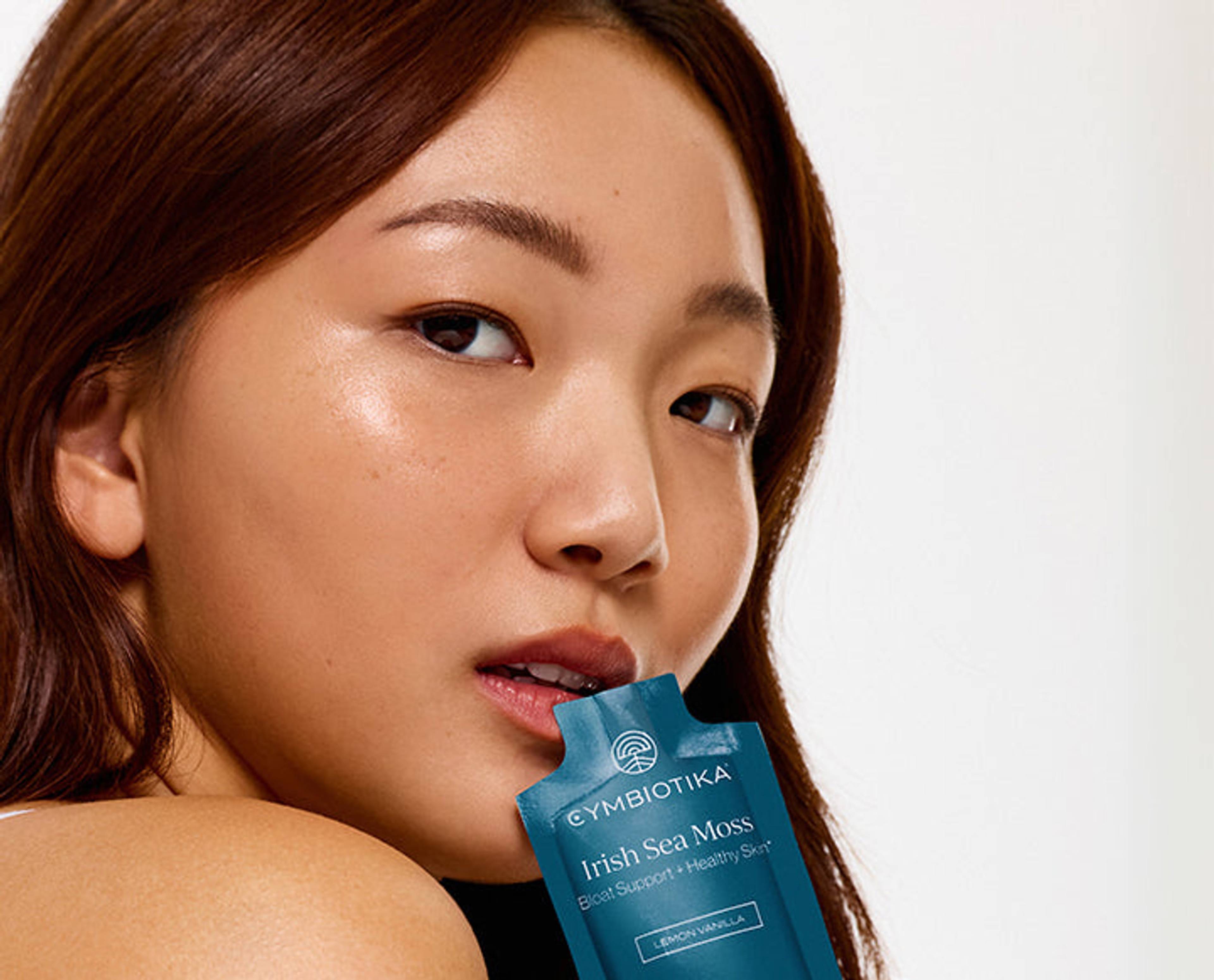 A woman poses with a blue Cymbiotika Irish Sea Moss sachet near her lips against a light background.