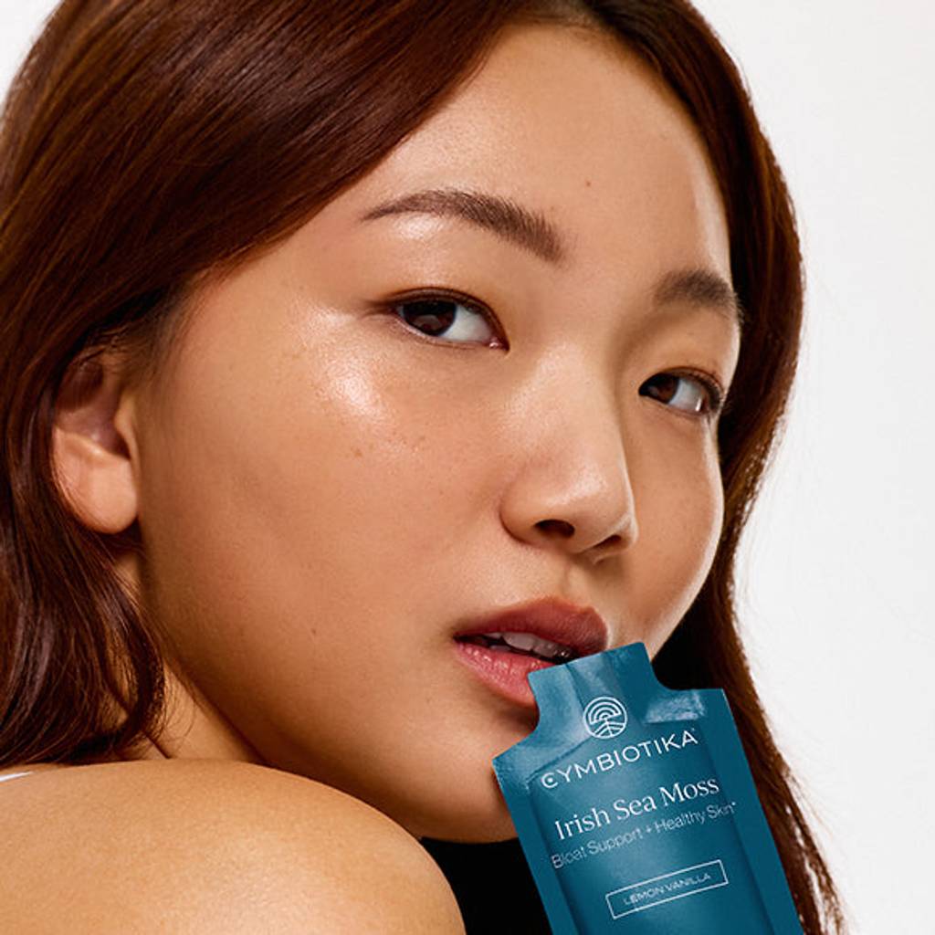 A woman poses with a blue Cymbiotika Irish Sea Moss sachet near her lips against a light background.