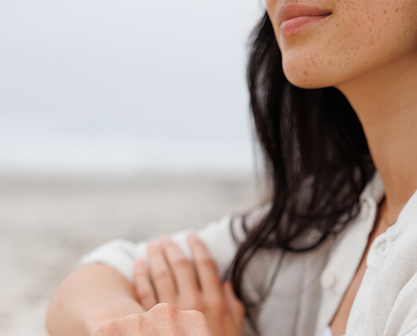 Close-up of a freckled woman with dark hair in a white shirt, arms folded outdoors.