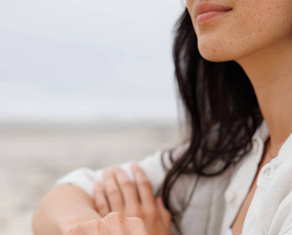 Close-up of a freckled woman with dark hair in a white shirt, arms folded outdoors.