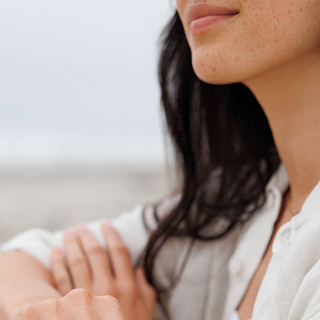 Close-up of a freckled woman with dark hair in a white shirt, arms folded outdoors.