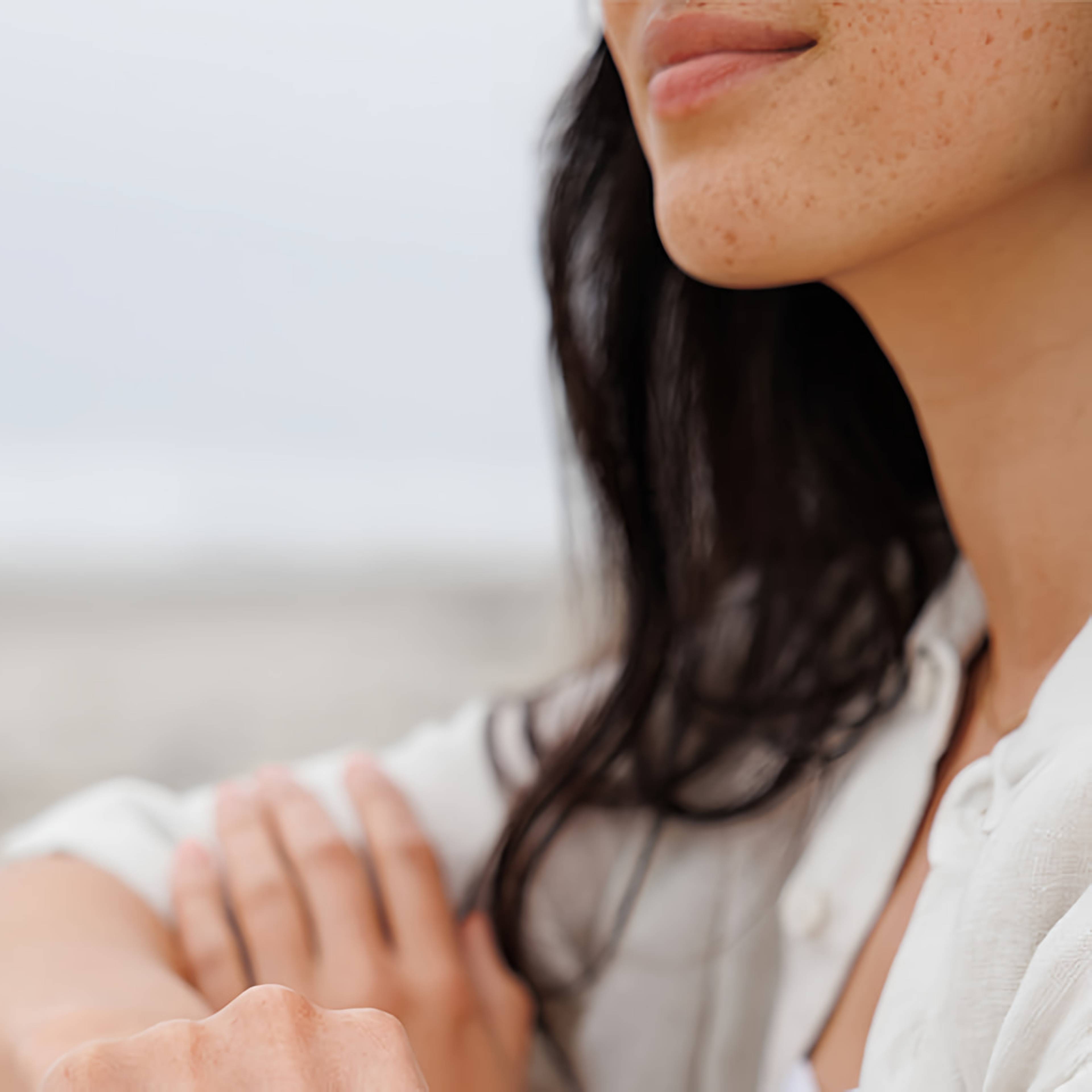 Close-up of a freckled woman with dark hair in a white shirt, arms folded outdoors.